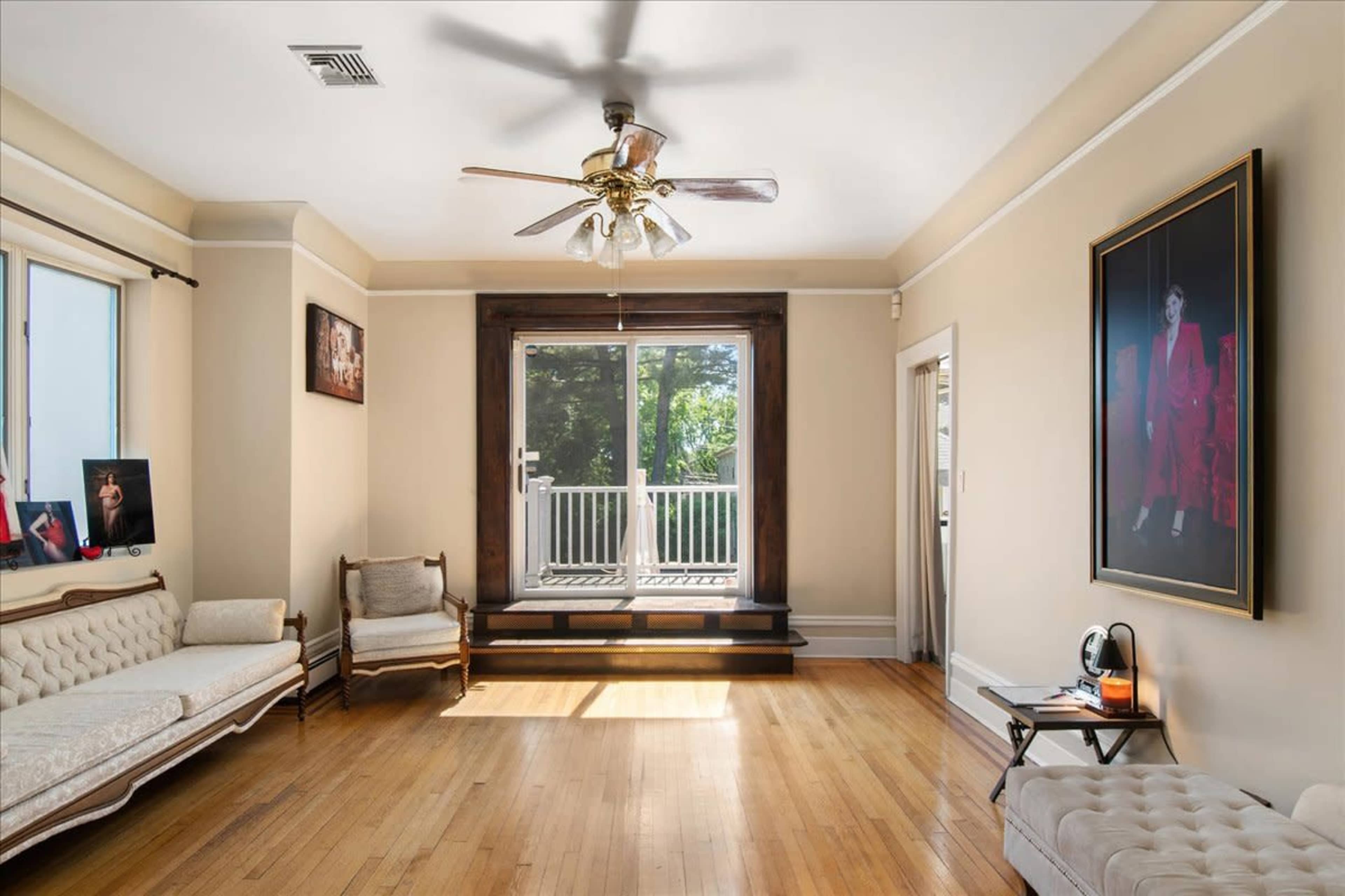 A living room with hardwood floors, a ceiling fan, and a large window leading to an outdoor deck, flanked by a sofa and a small armchair.