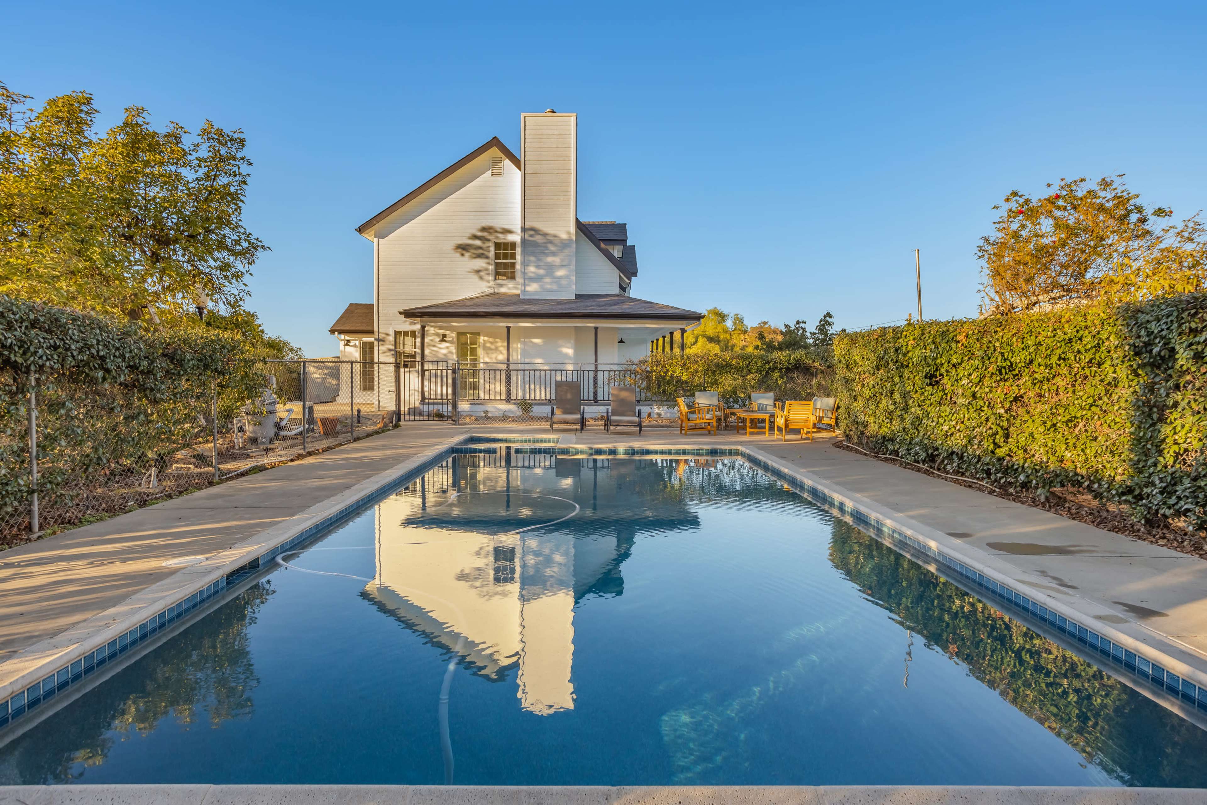 A clear blue swimming pool reflects a white house surrounded by green hedges and trees.
