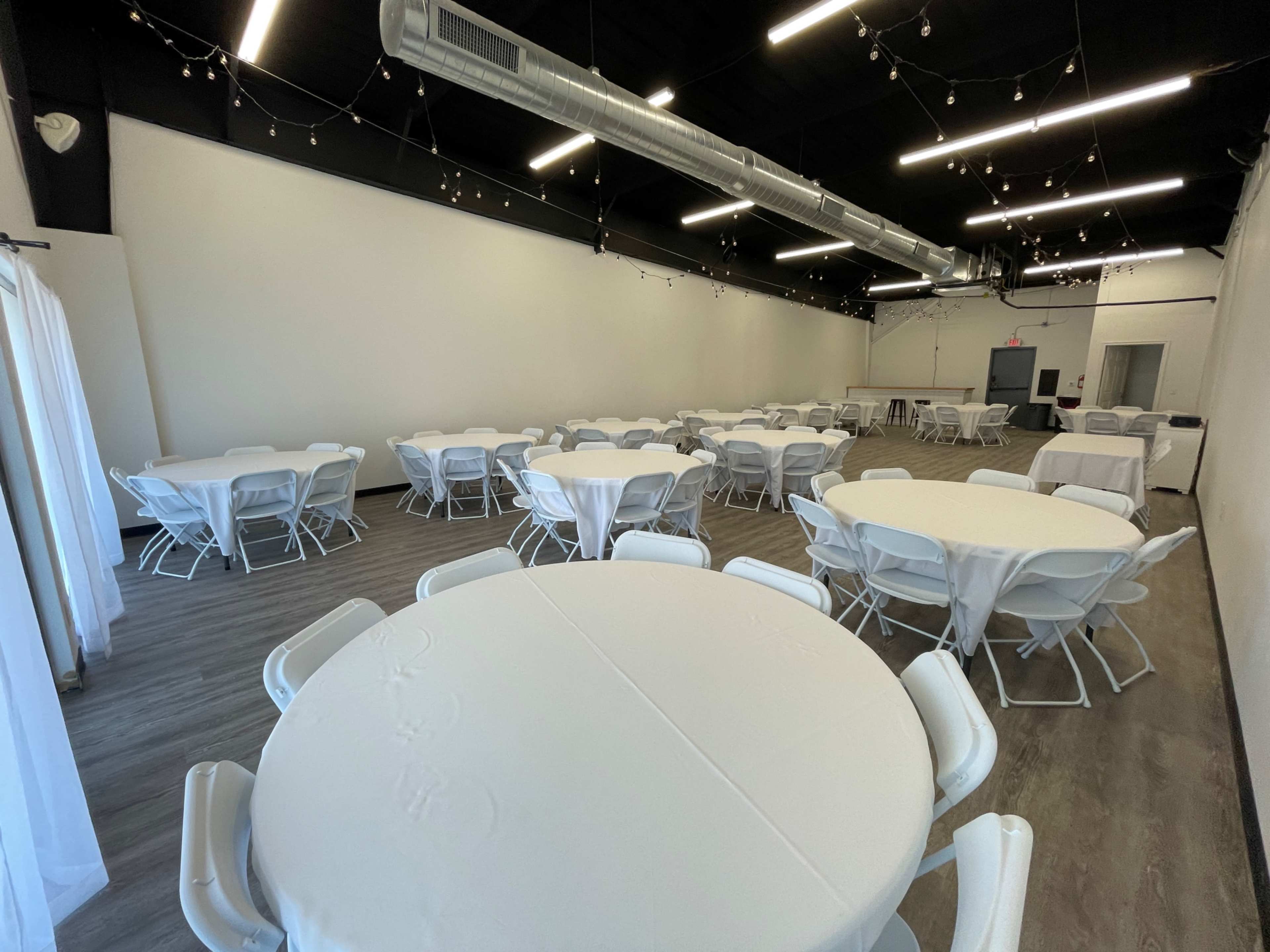 An empty event space with several round tables covered in white tablecloths arranged in a large room with overhead string lights.
