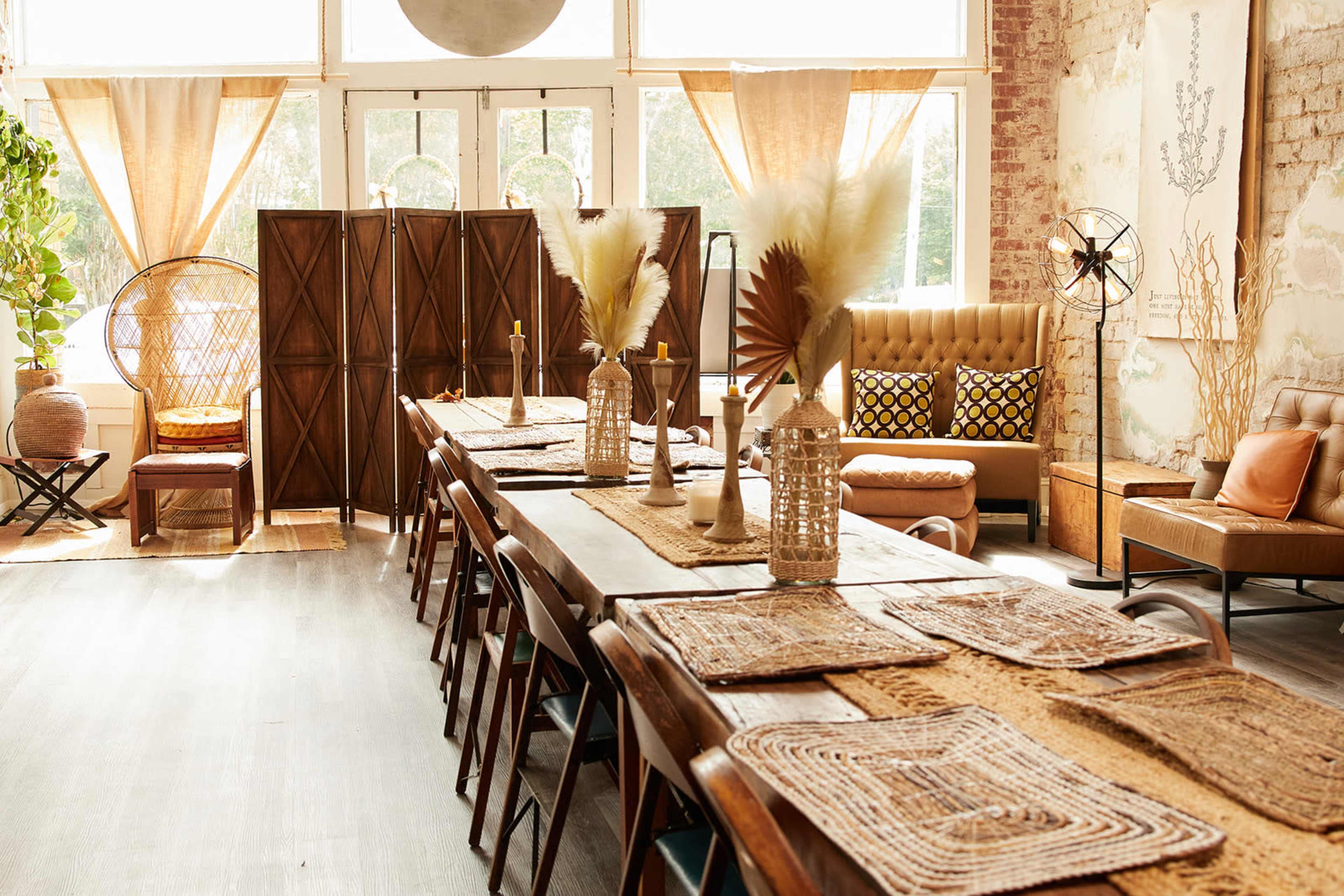 A spacious dining area with a long wooden table set for a meal, surrounded by minimalist decor, including plants, woven elements, and a folding wooden screen.
