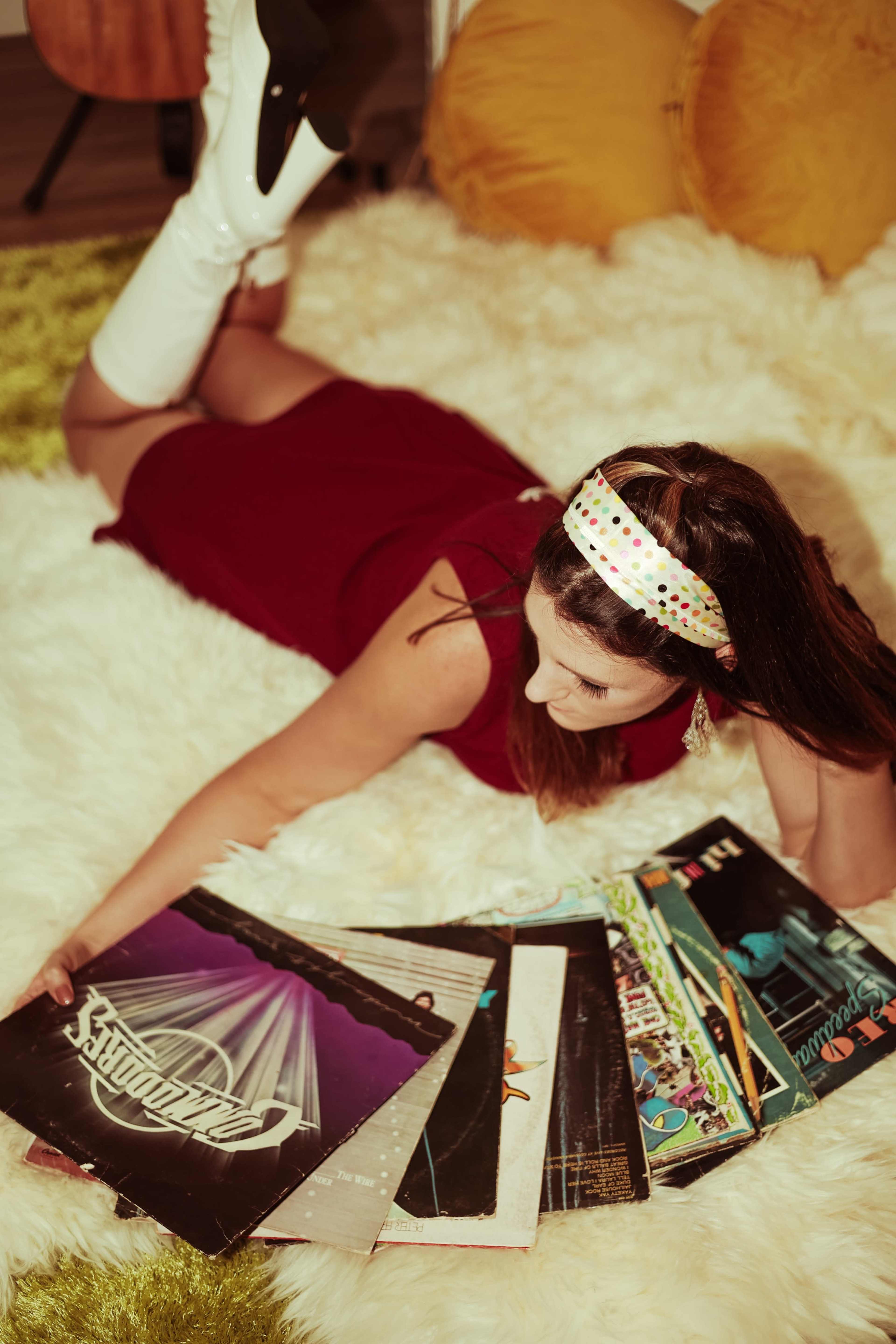 A woman in a red dress and white knee-high boots lies on a shaggy rug while browsing through a collection of vinyl records.