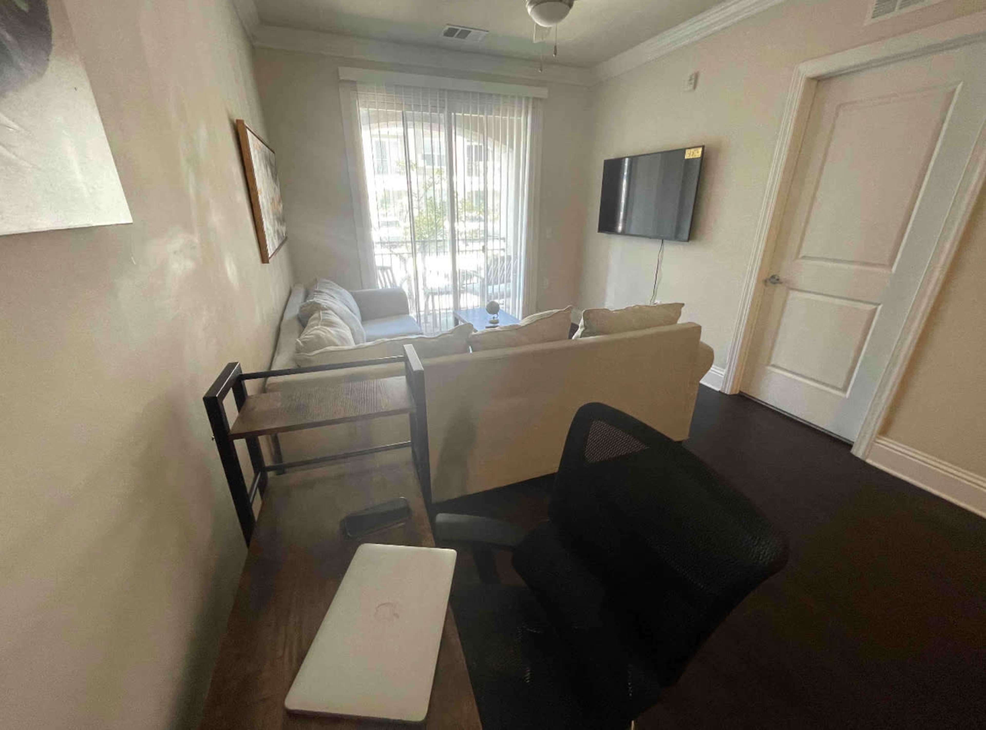 A small living area features a couch, a desk with a laptop, and a television mounted on the wall, with natural light streaming through the window.