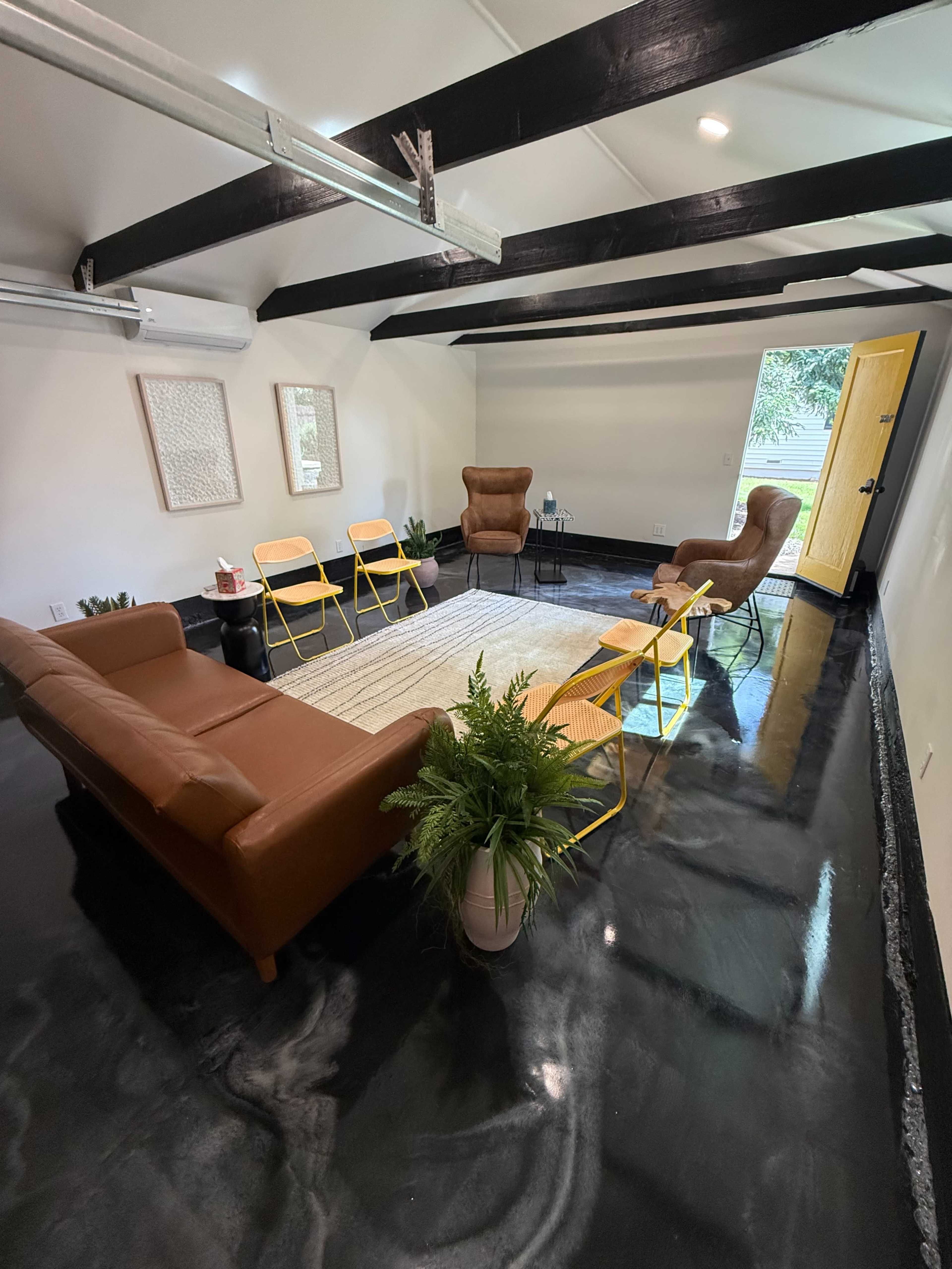 The image shows a modern room with a glossy black floor, featuring a brown leather sofa, two armchairs, and yellow folding chairs arranged around a central area rug.