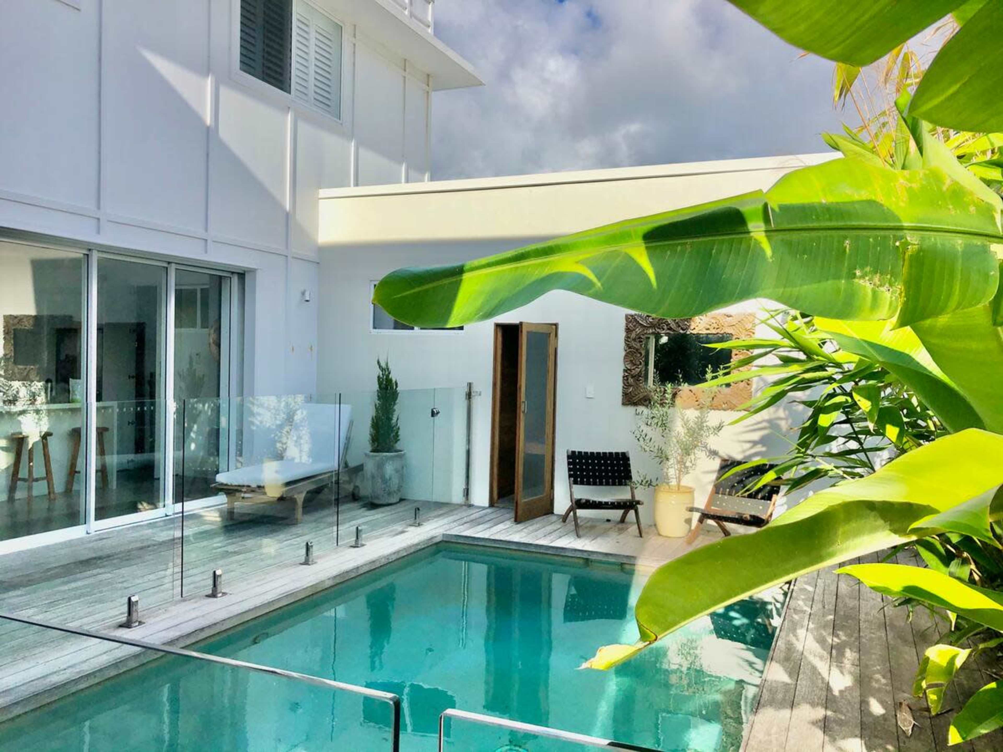 A modern villa features a swimming pool surrounded by wooden decking and lush tropical plants.