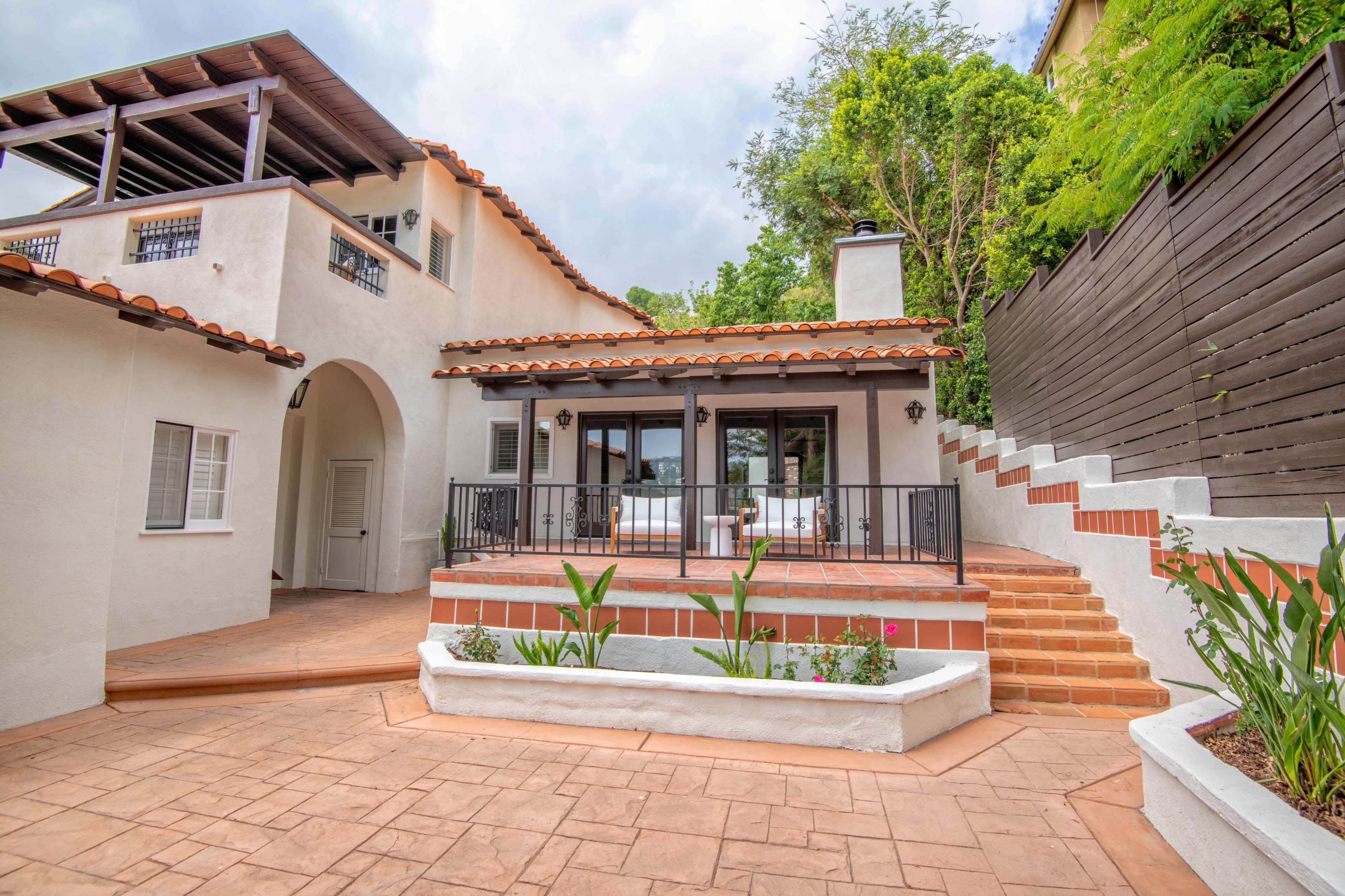 A two-story house with a terracotta roof, a covered balcony, and a landscaped outdoor terrace leading to a set of stairs.