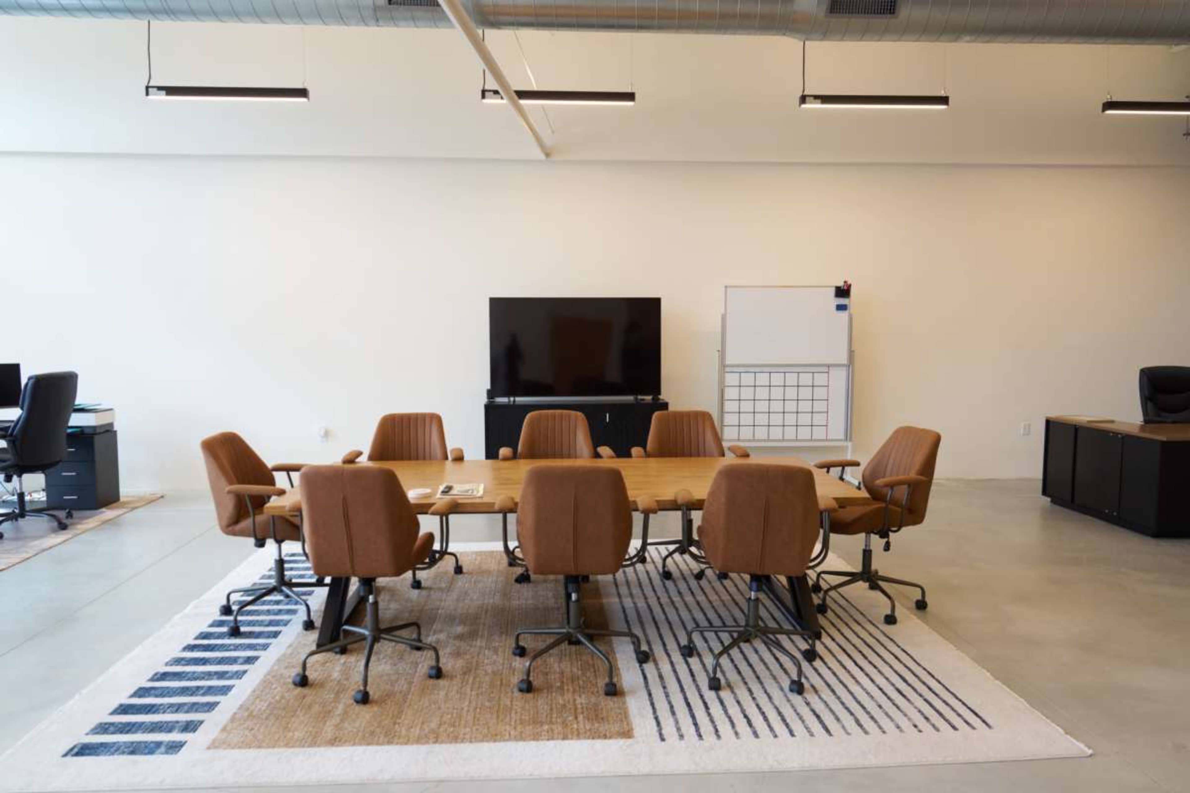 The image shows a conference room with a large wooden table surrounded by eight brown rolling chairs, a television on the wall, and a whiteboard nearby.