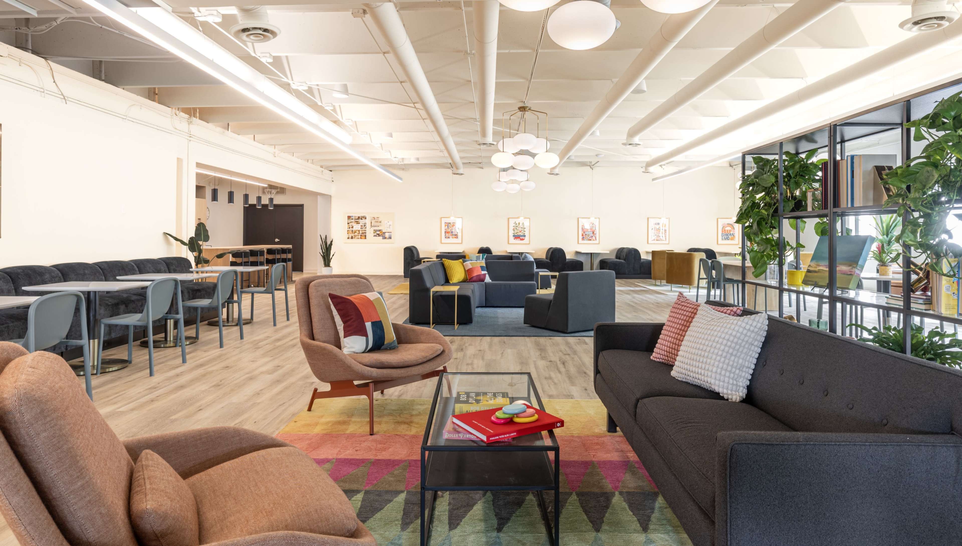The image shows a spacious, modern lounge area with a variety of seating arrangements, including sofas and armchairs, surrounded by plants and colorful decor.