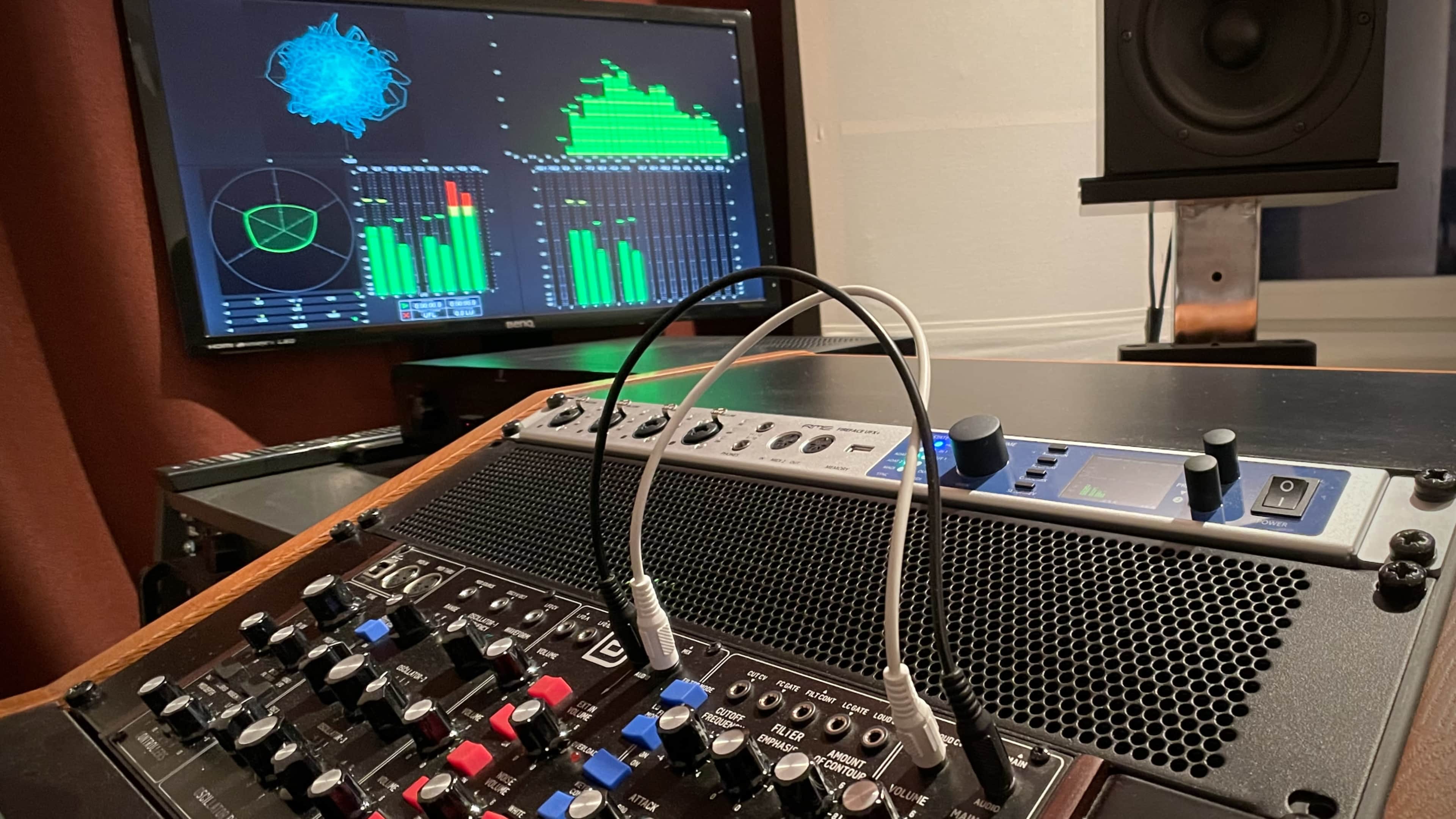 A digital audio workstation setup features an audio interface with various controls and two connected cables, alongside a monitor displaying sound visualizations and audio levels.