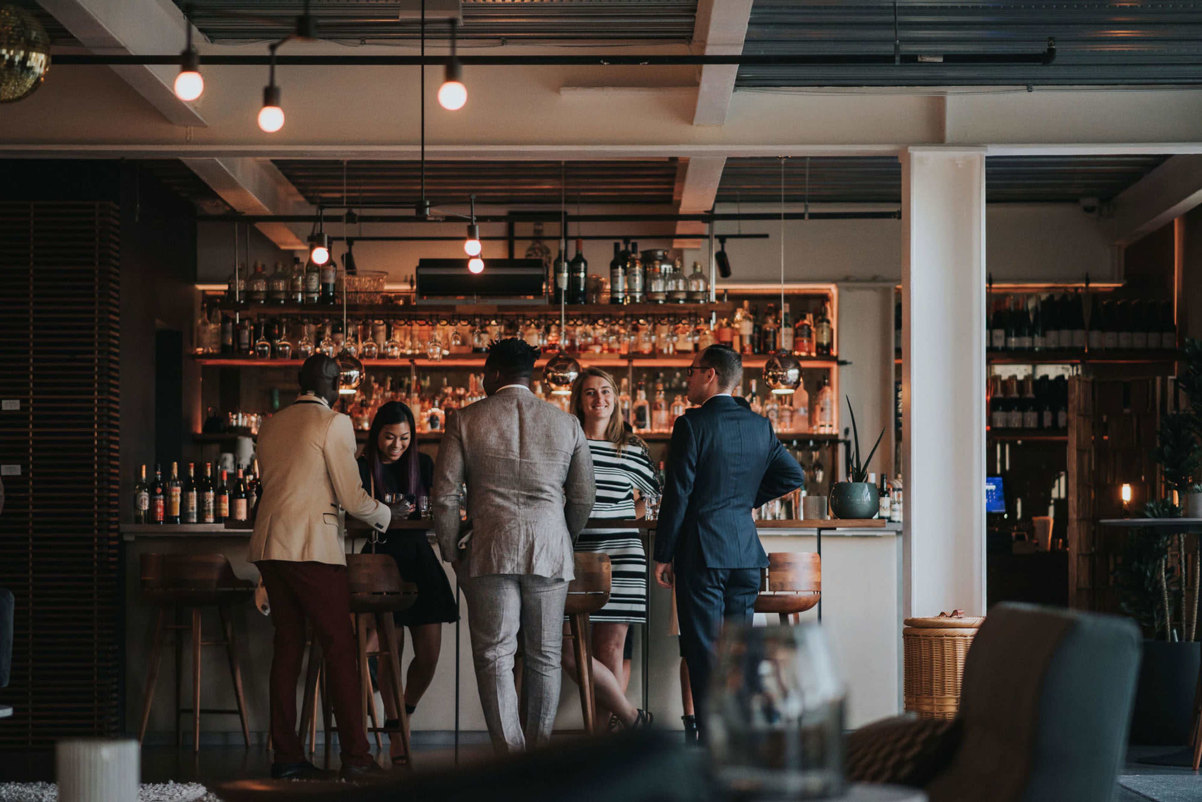 A group of six people in formal attire are gathered around a bar in a well-lit lounge, with shelves of bottles visible in the background.