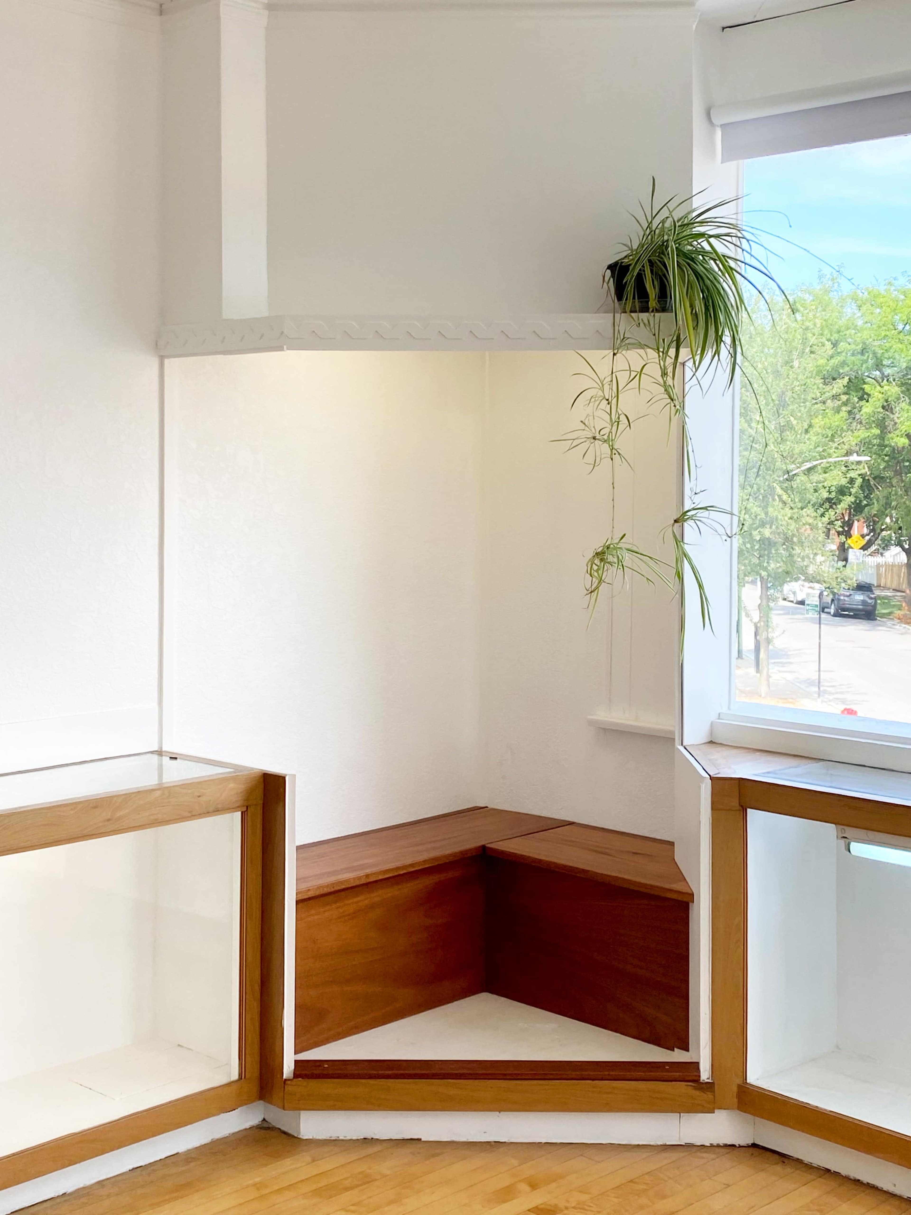 The image shows a bright, minimalistic corner of a room with a wooden bench and a window featuring a plant.