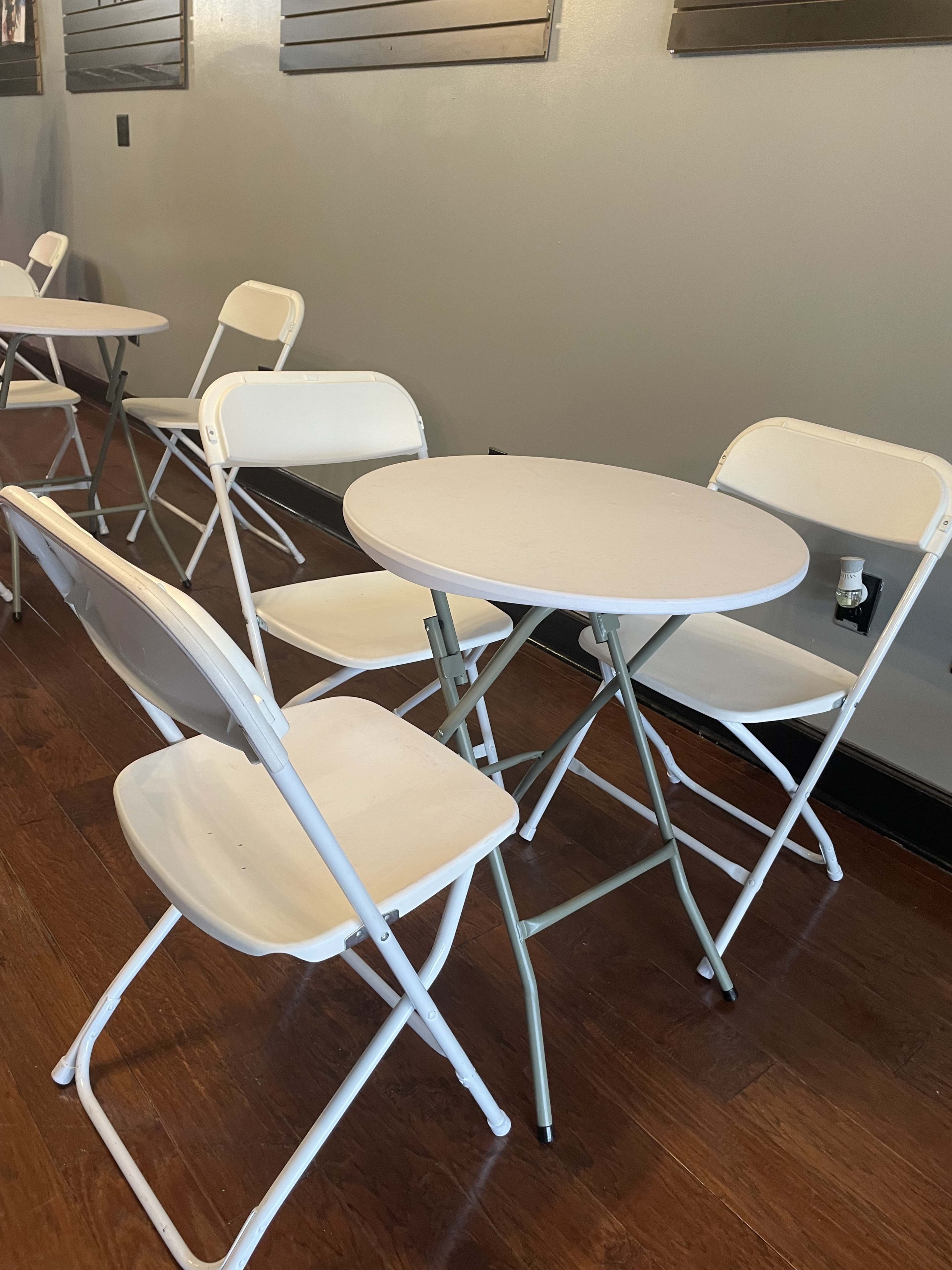 A small round table is surrounded by four white folding chairs in a room with wooden flooring.