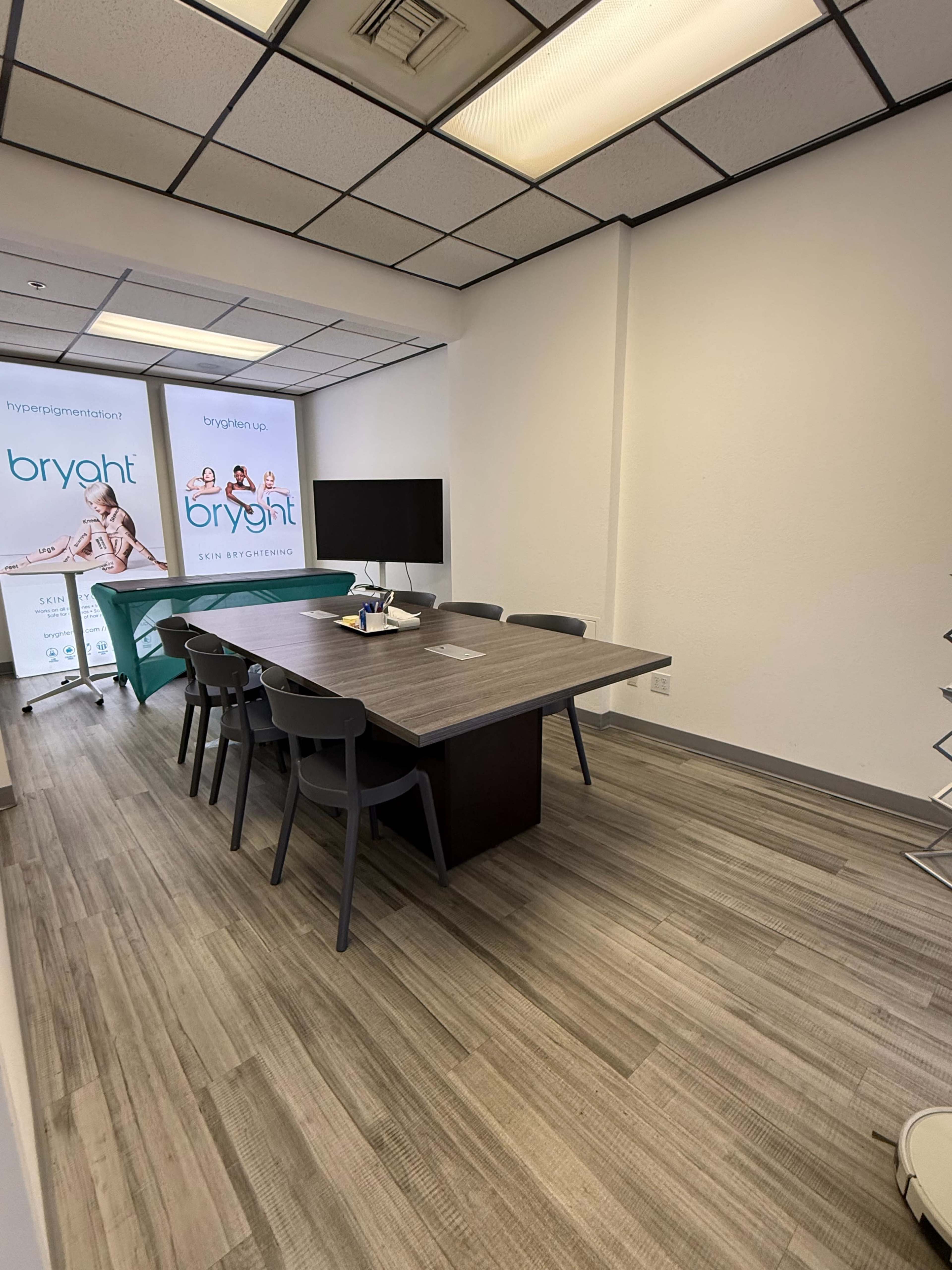 The image shows a modern conference room with a large wooden table surrounded by gray chairs, a flat-screen monitor, and branding banners on the wall.