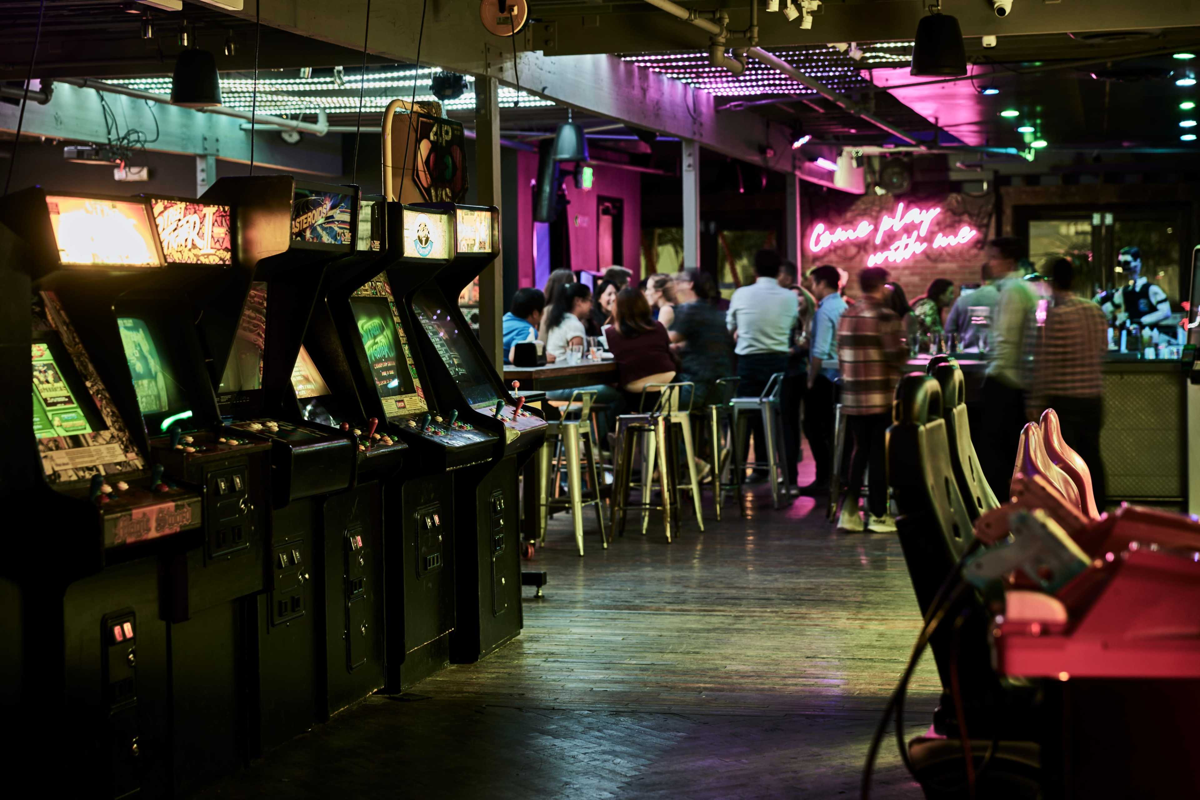 A dimly lit arcade with several vintage arcade machines on the left and a bar area filled with patrons in the background.