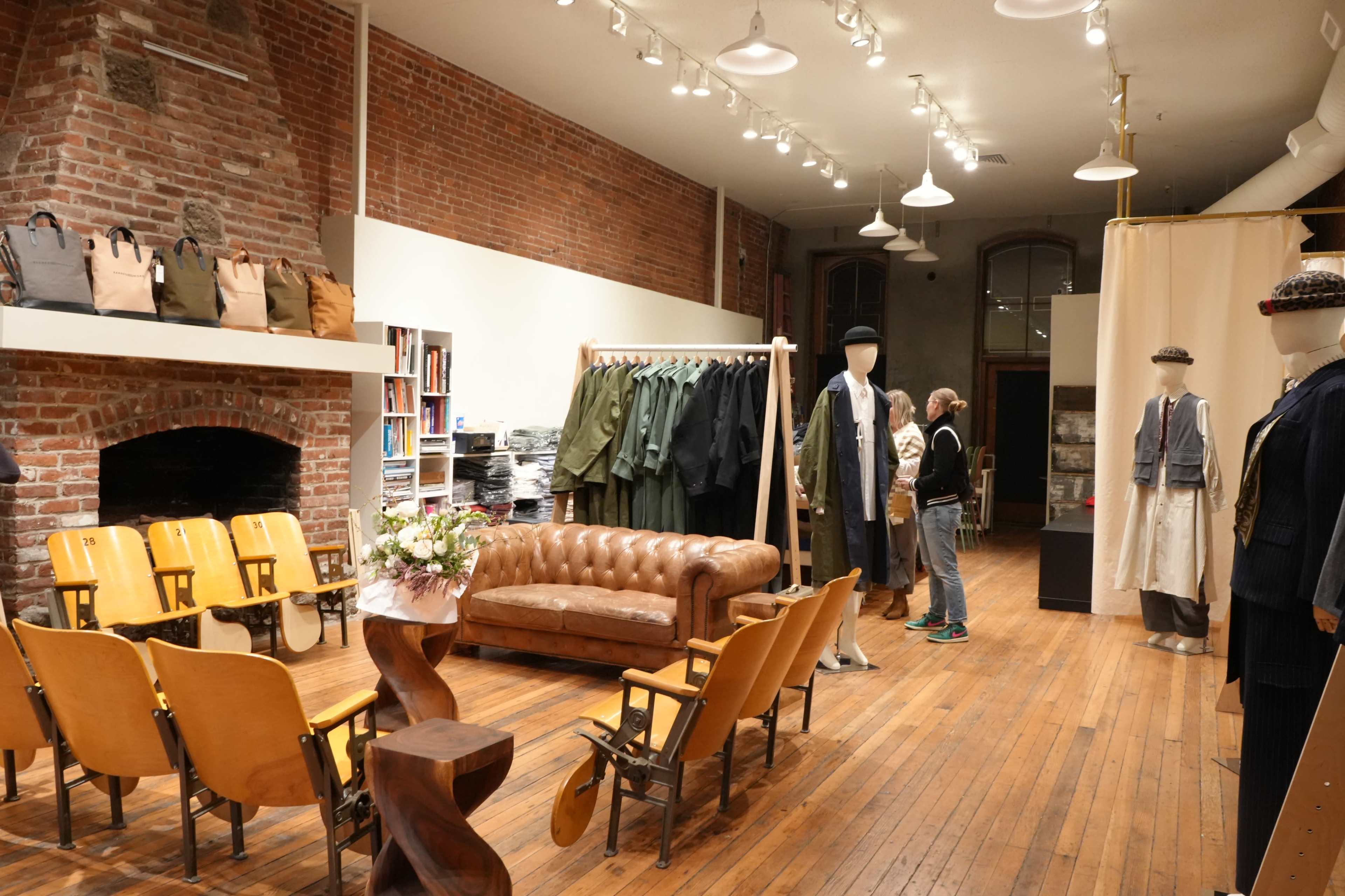 The image shows a spacious clothing store with brick walls, wooden floors, and a mix of hanging garments and displayed mannequins, alongside a vintage leather sofa and wooden seating.