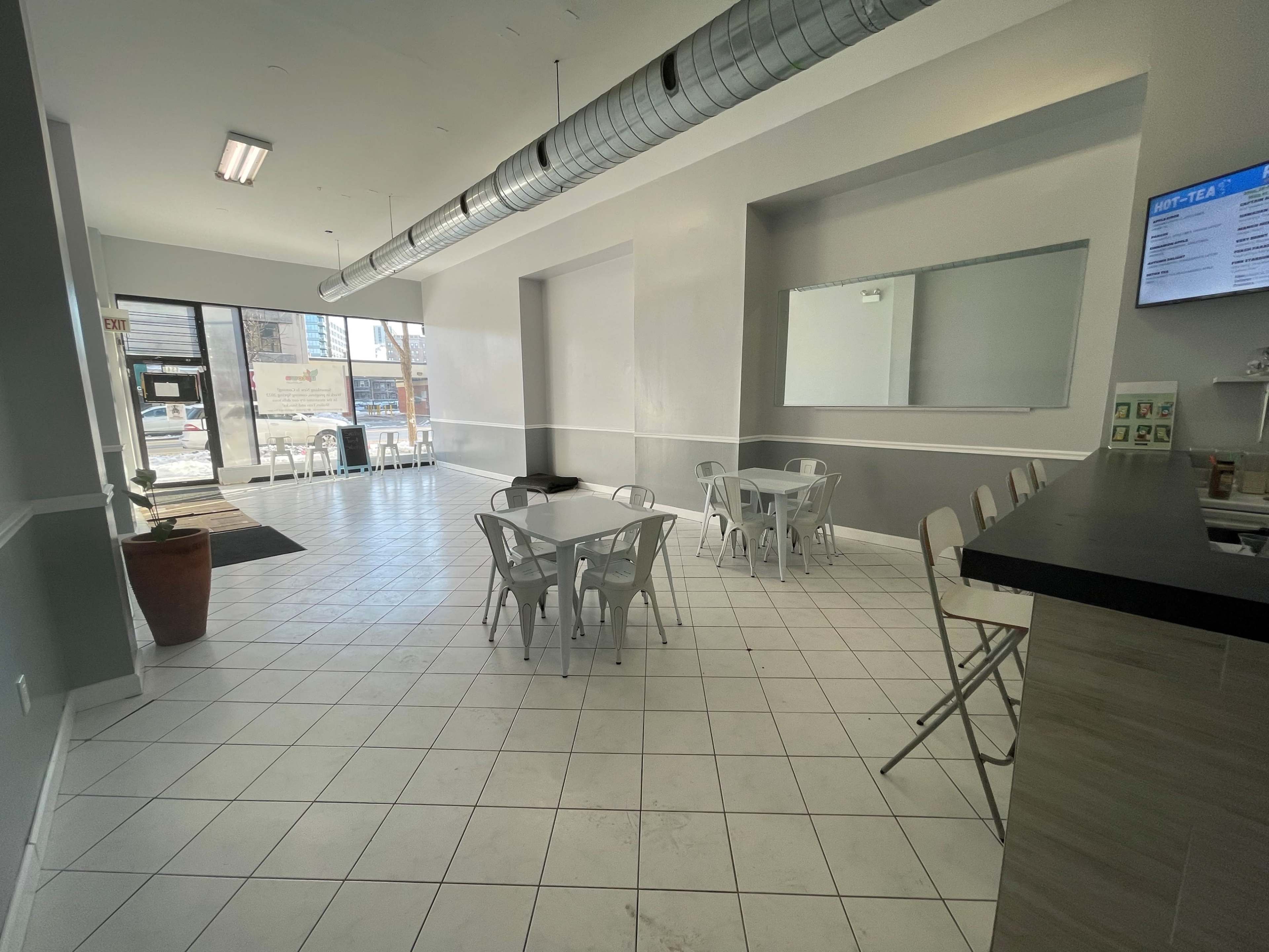 A modern, empty café with white walls, tiled flooring, and metal tables and chairs, featuring a large window at the entrance.