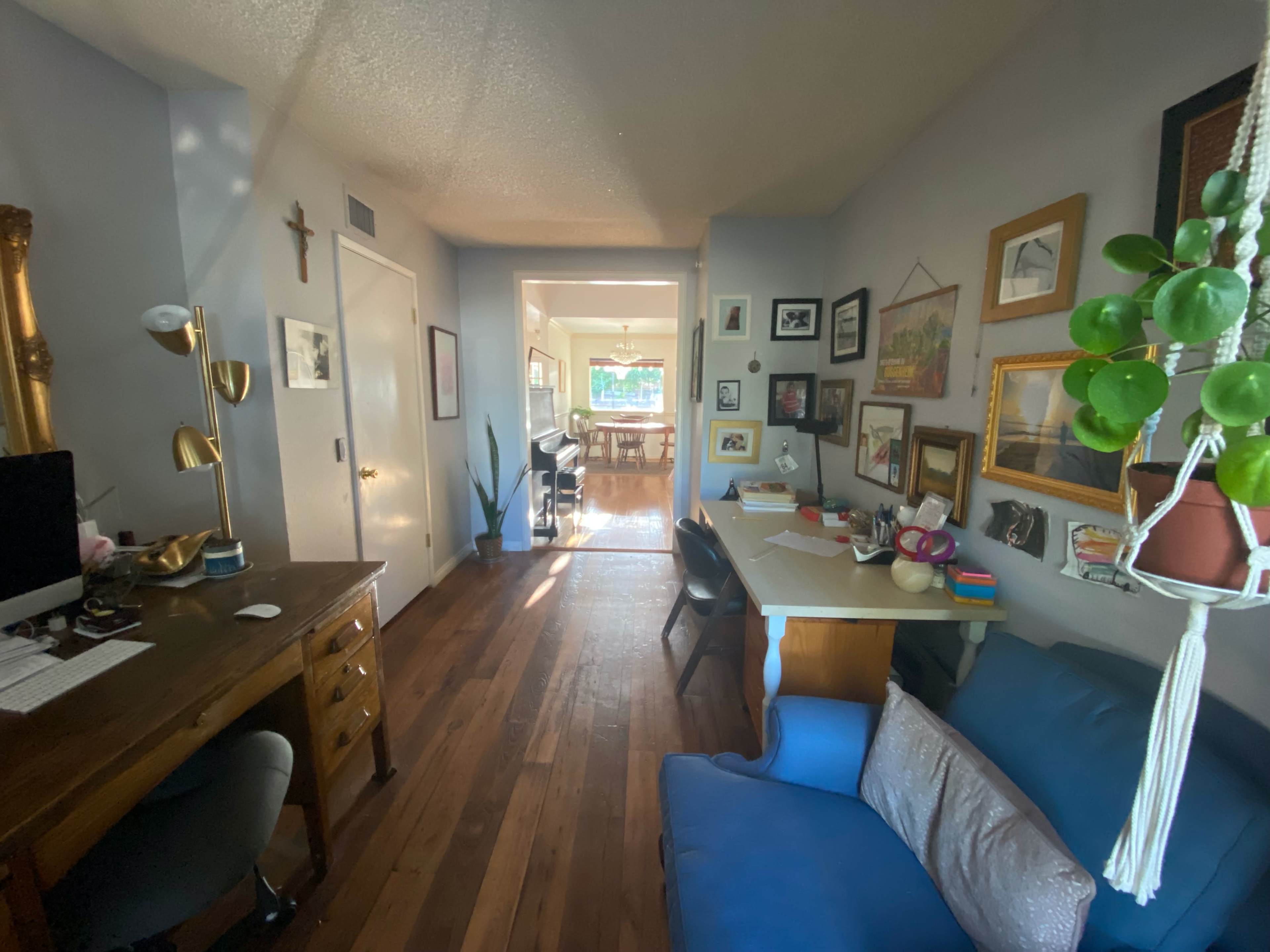 A hallway leads from a small desk area to a well-lit dining space, with framed art on the walls and a blue sofa nearby.
