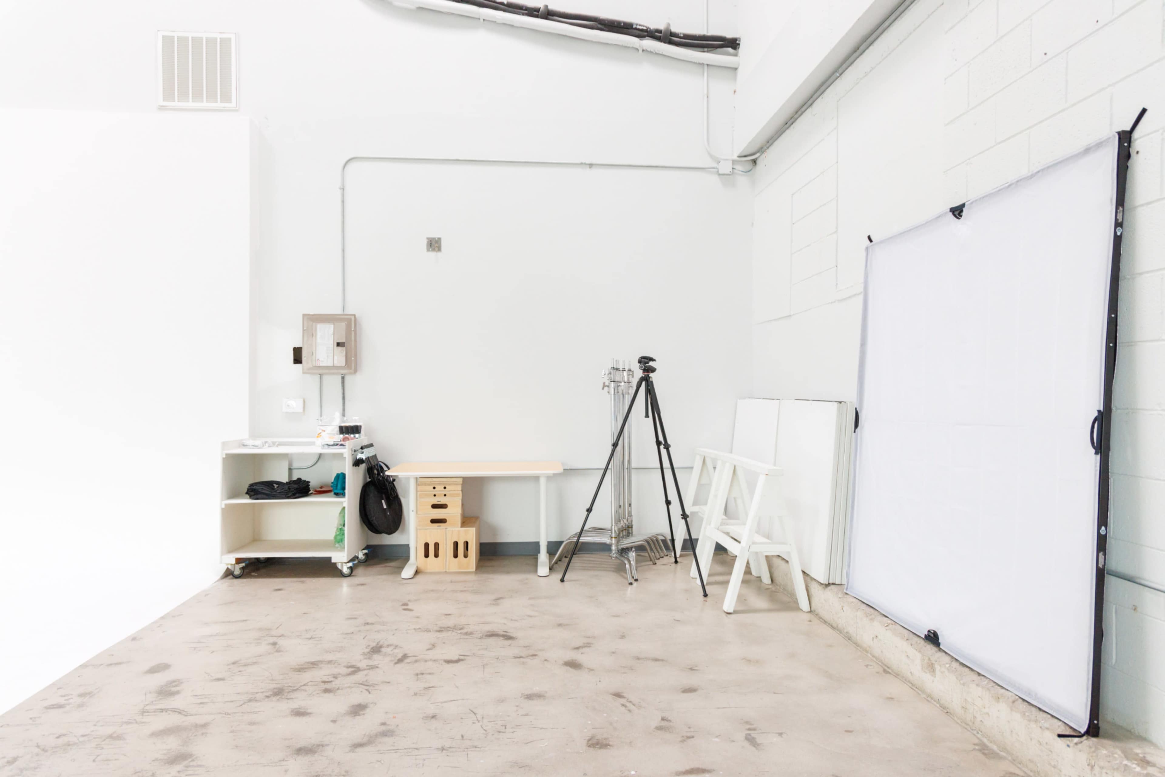 The image shows a minimalistic photo studio interior featuring a tripod, a work table, a light reflector, and bare concrete flooring.