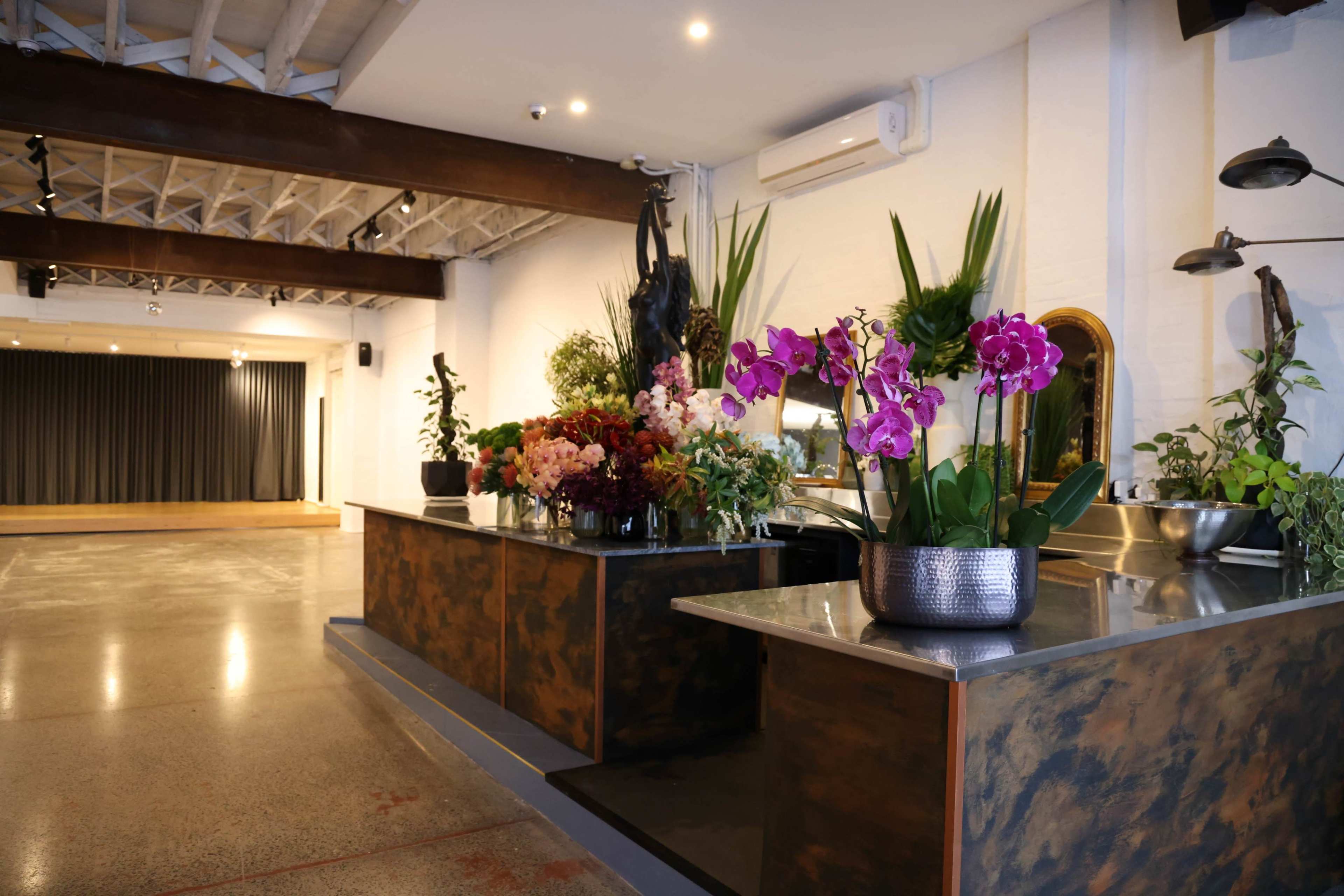 A spacious floral shop with a counter displaying various floral arrangements and plants, set against a minimalist interior featuring wooden beams and a stage area in the background.
