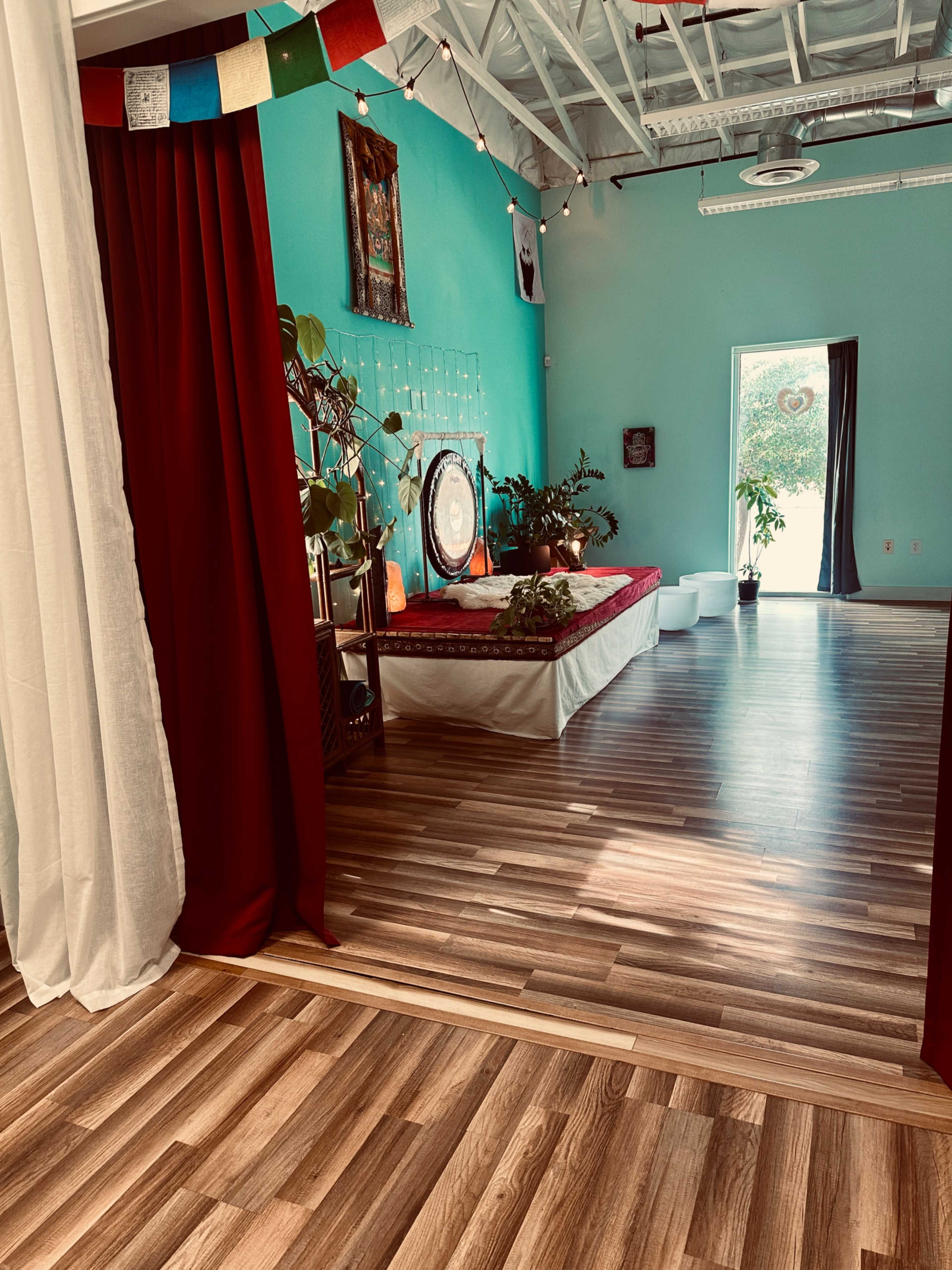 The image shows a brightly colored room featuring wooden flooring, a red draped area, and a meditation space with plants, decorations, and a large circular object.