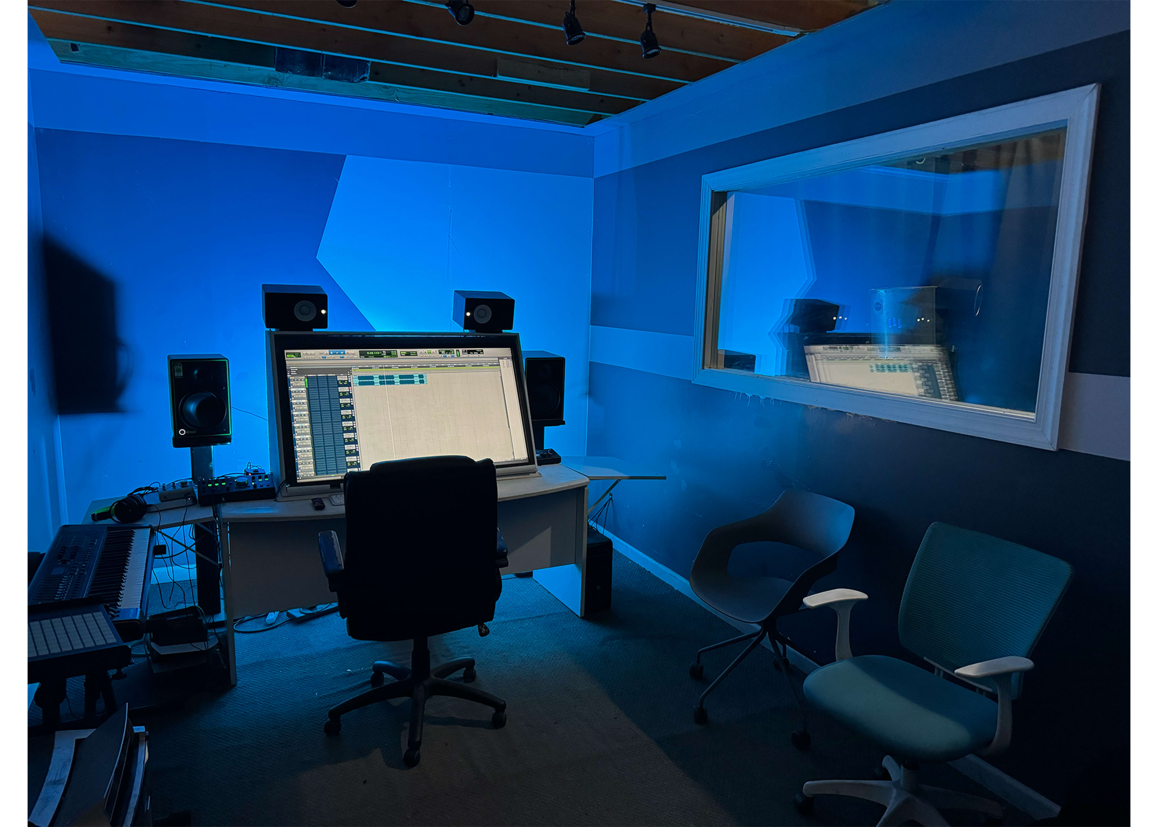 The image shows a music production studio with a large monitor, speakers, and chairs set against a blue-lit wall.