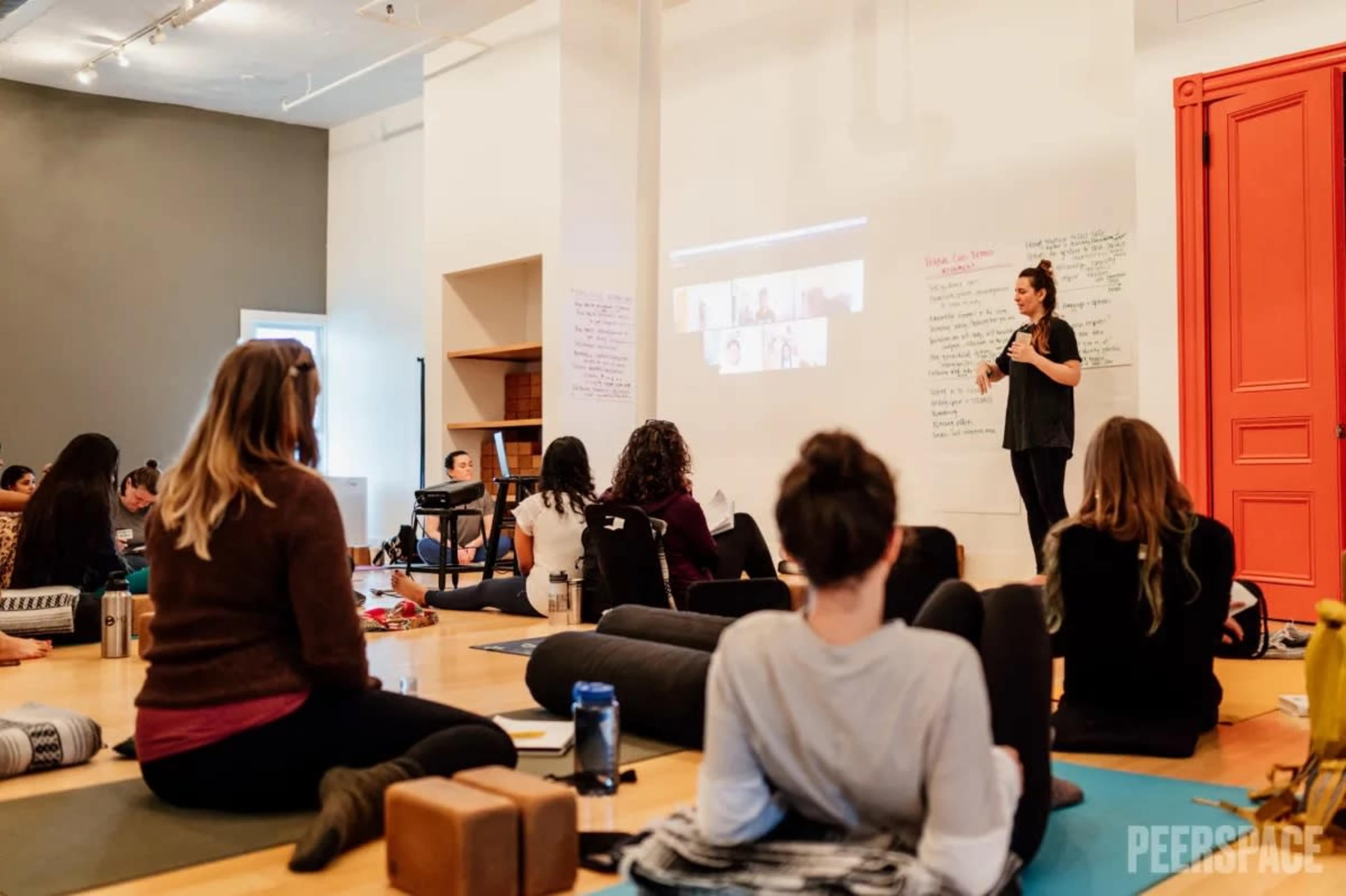 A group of participants sits on yoga mats in a studio while a speaker presents in front of them with a projected screen.