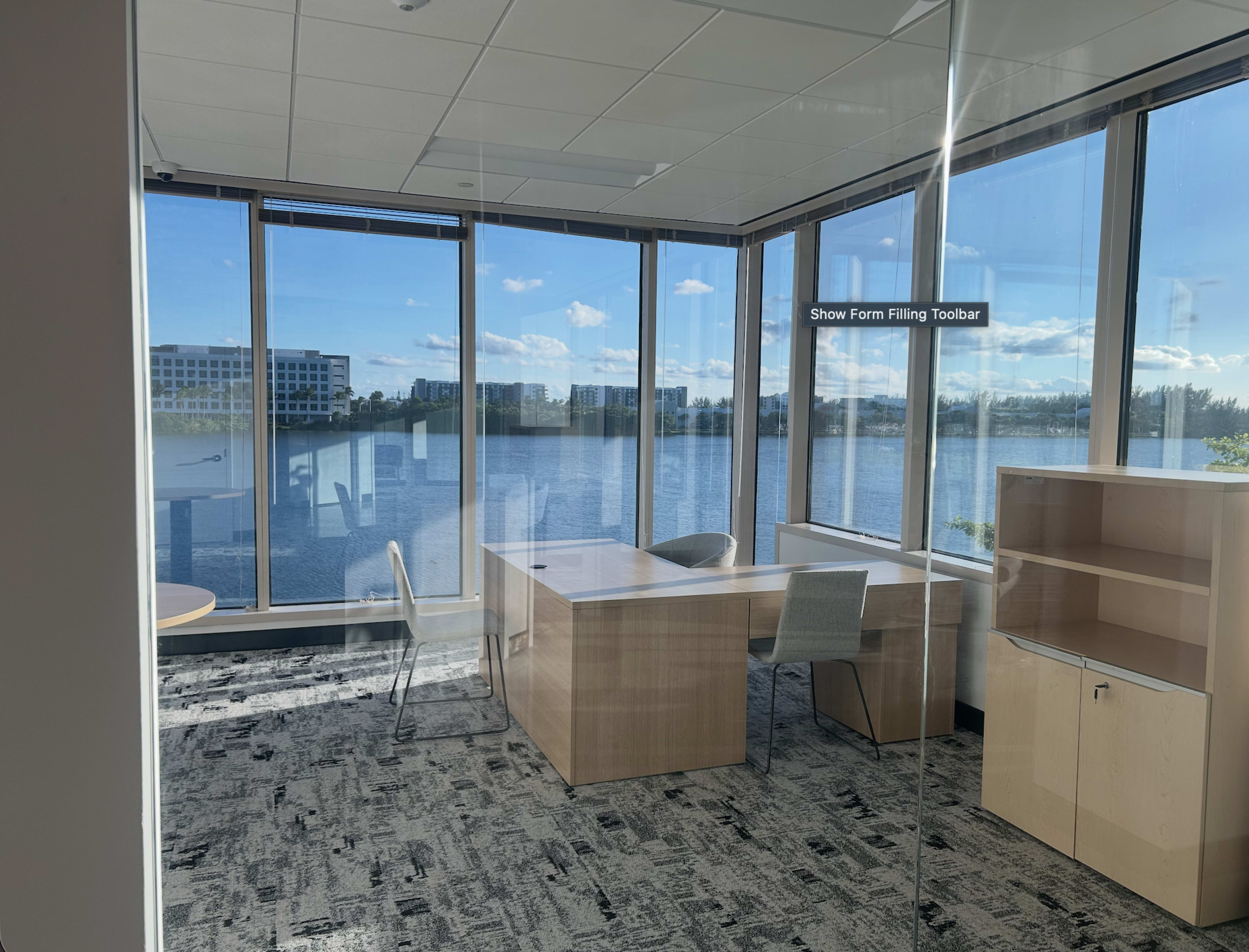 An office space features a desk and chair with large windows overlooking a body of water and distant buildings.