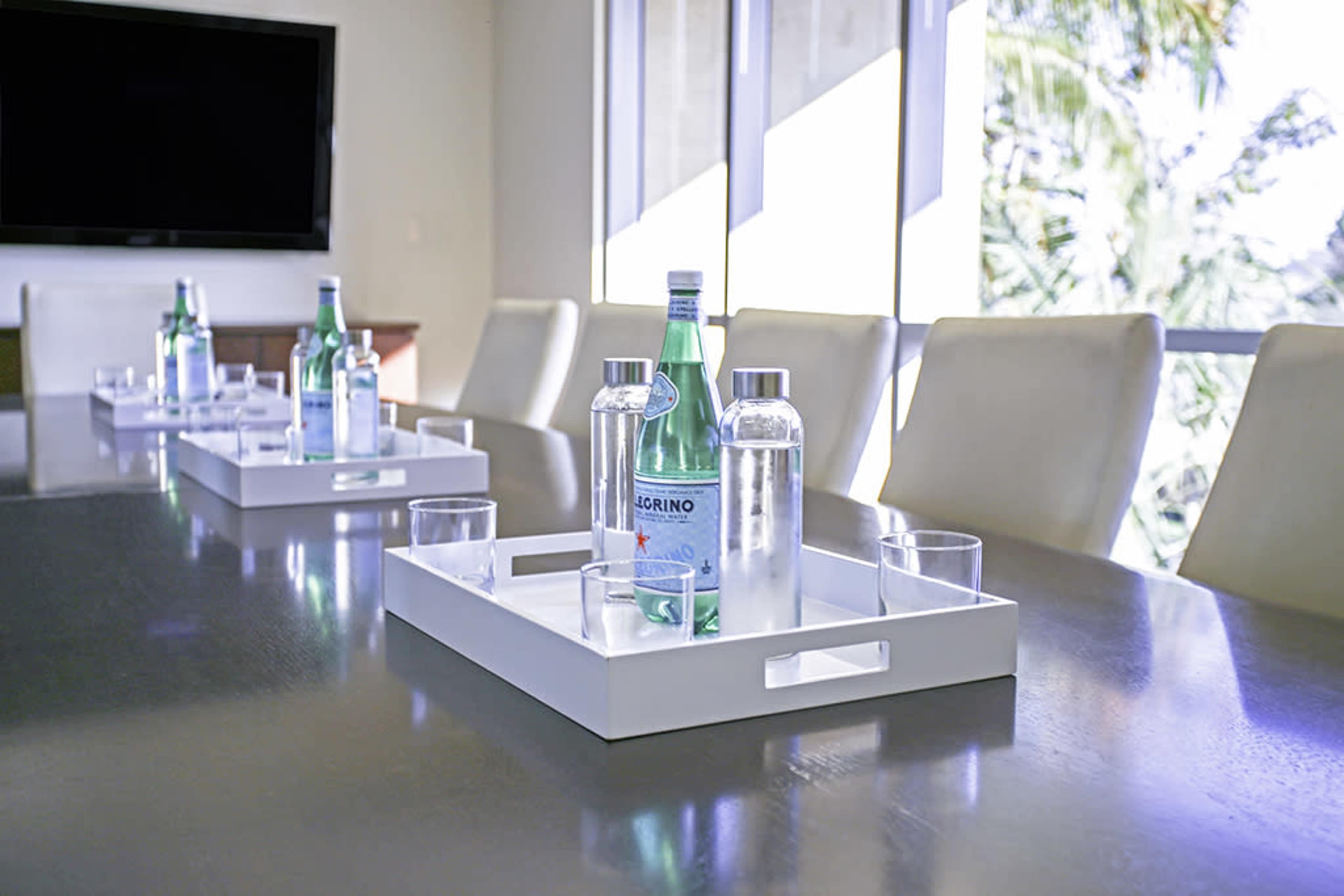 A conference table is set with trays containing bottles of sparkling water and glasses, with large windows in the background.