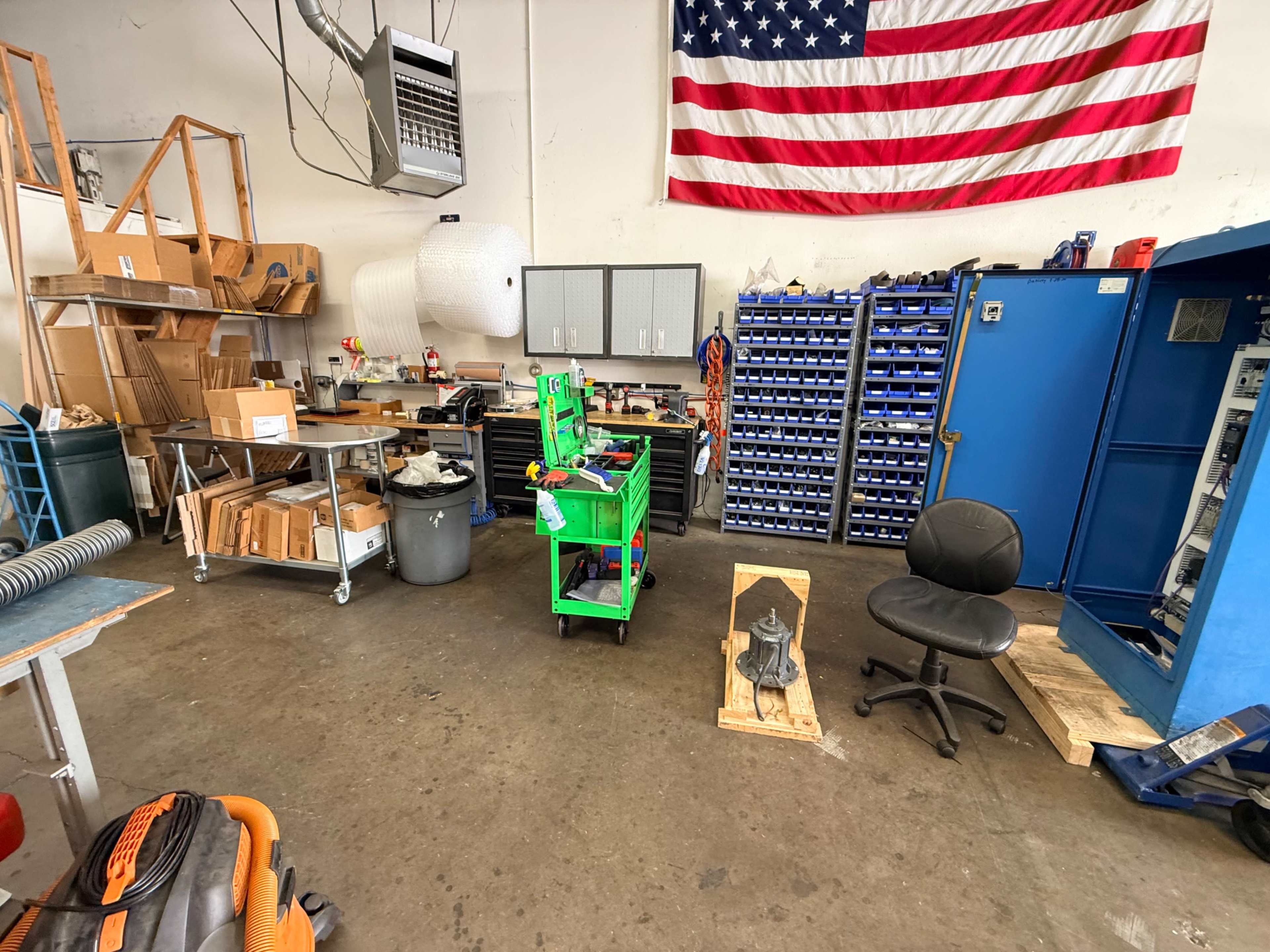 The image shows an industrial workspace with various tools and materials, a flag hanging on the wall, and a cart in the center of the room.