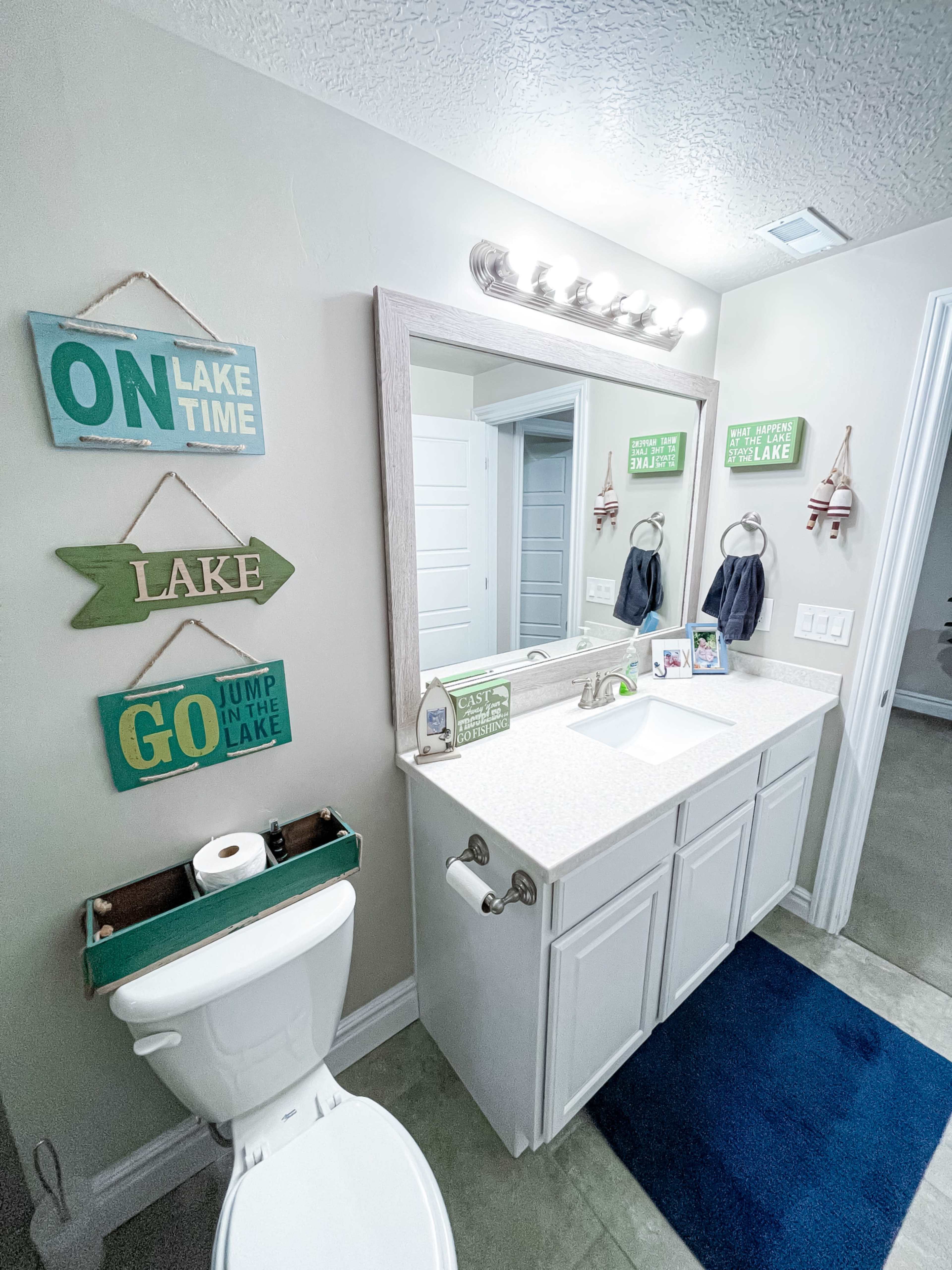 A bathroom features a light-colored vanity with a large mirror above, flanked by various wall signs, and a toilet positioned beside it.