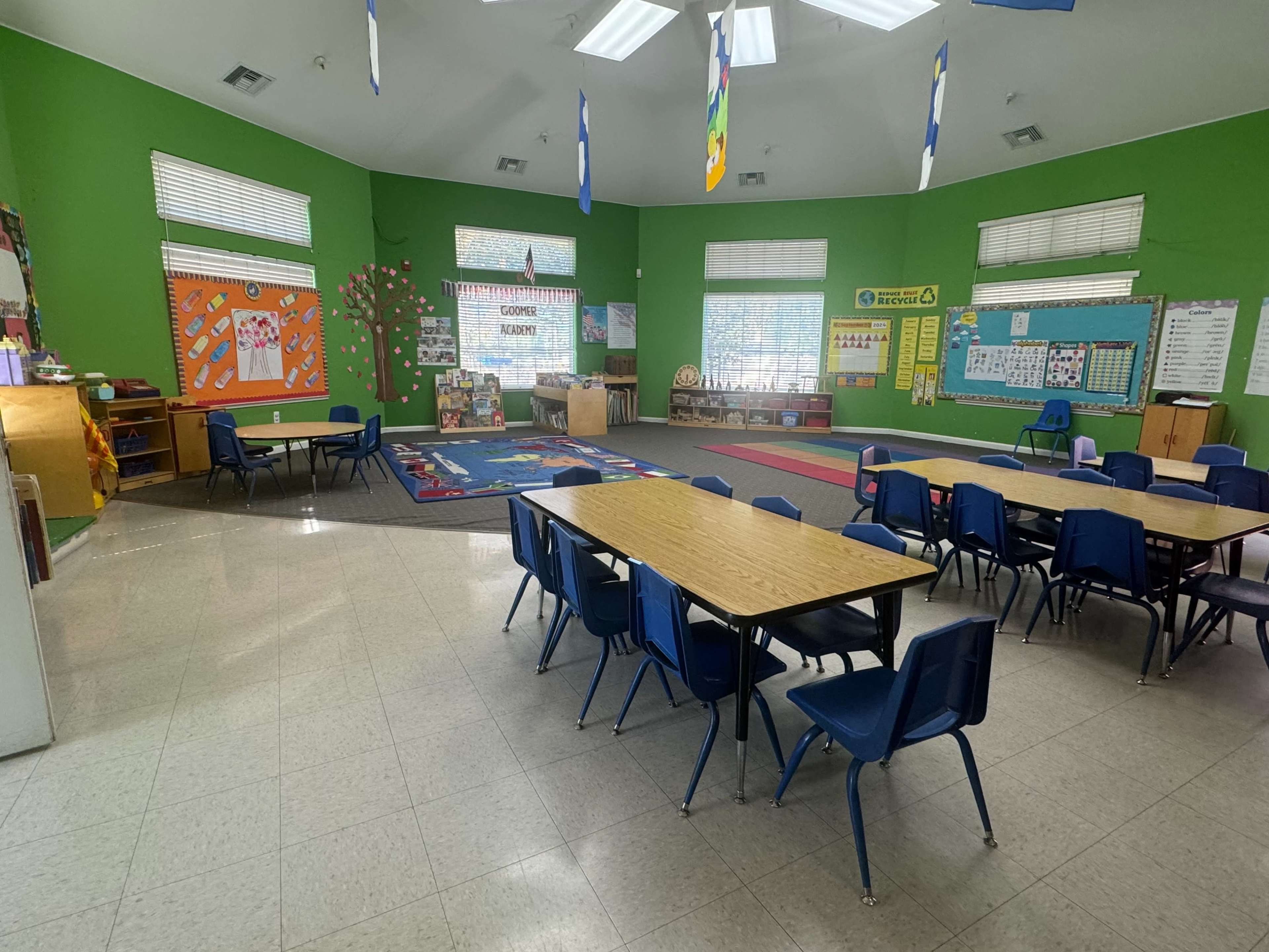 The image shows a classroom with green walls, wooden tables, and blue chairs arranged around the space, featuring educational posters and a carpeted area for activities.