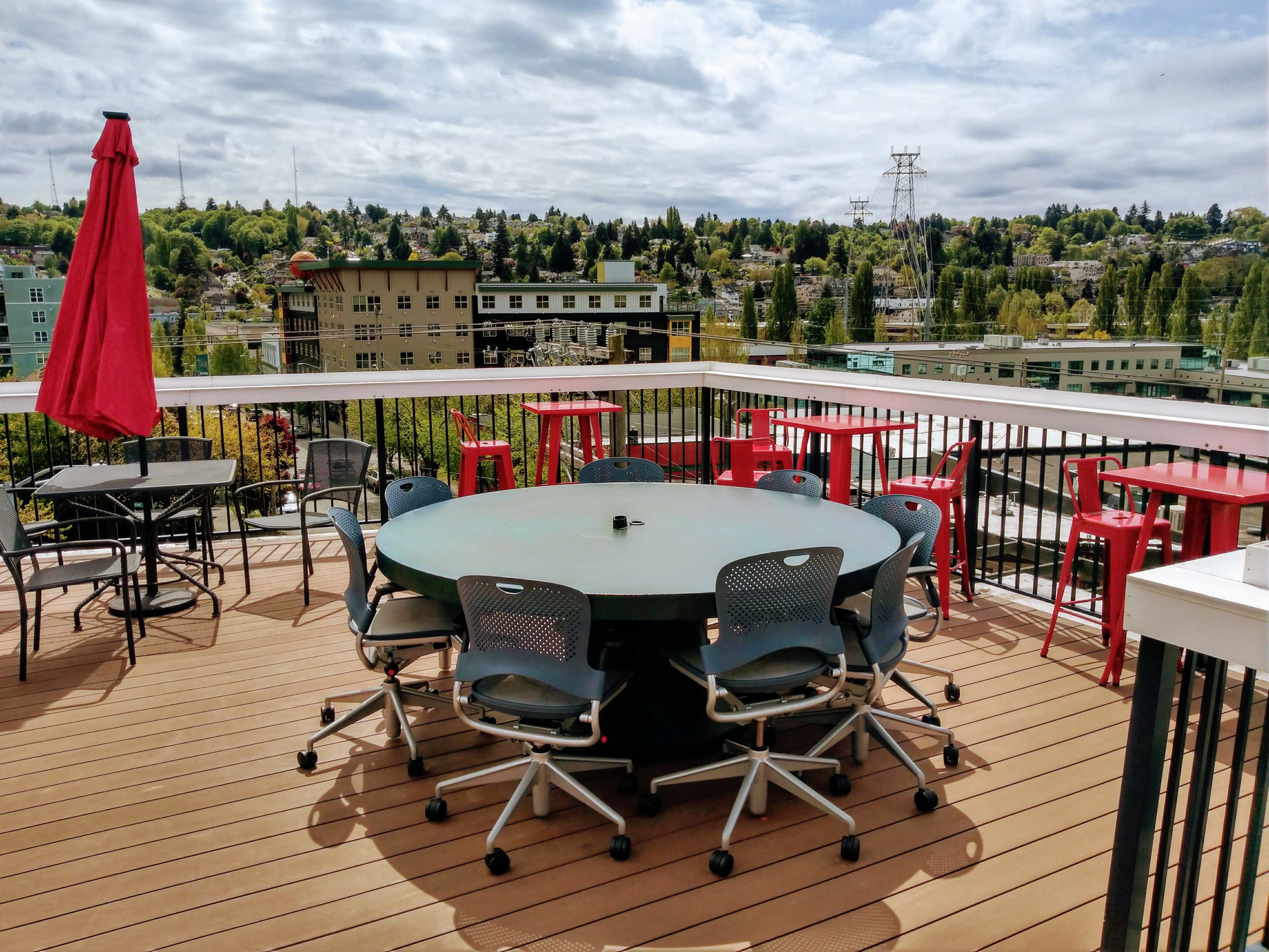 A rooftop terrace with a round table surrounded by chairs, a red umbrella, and a view of buildings and trees in the background.