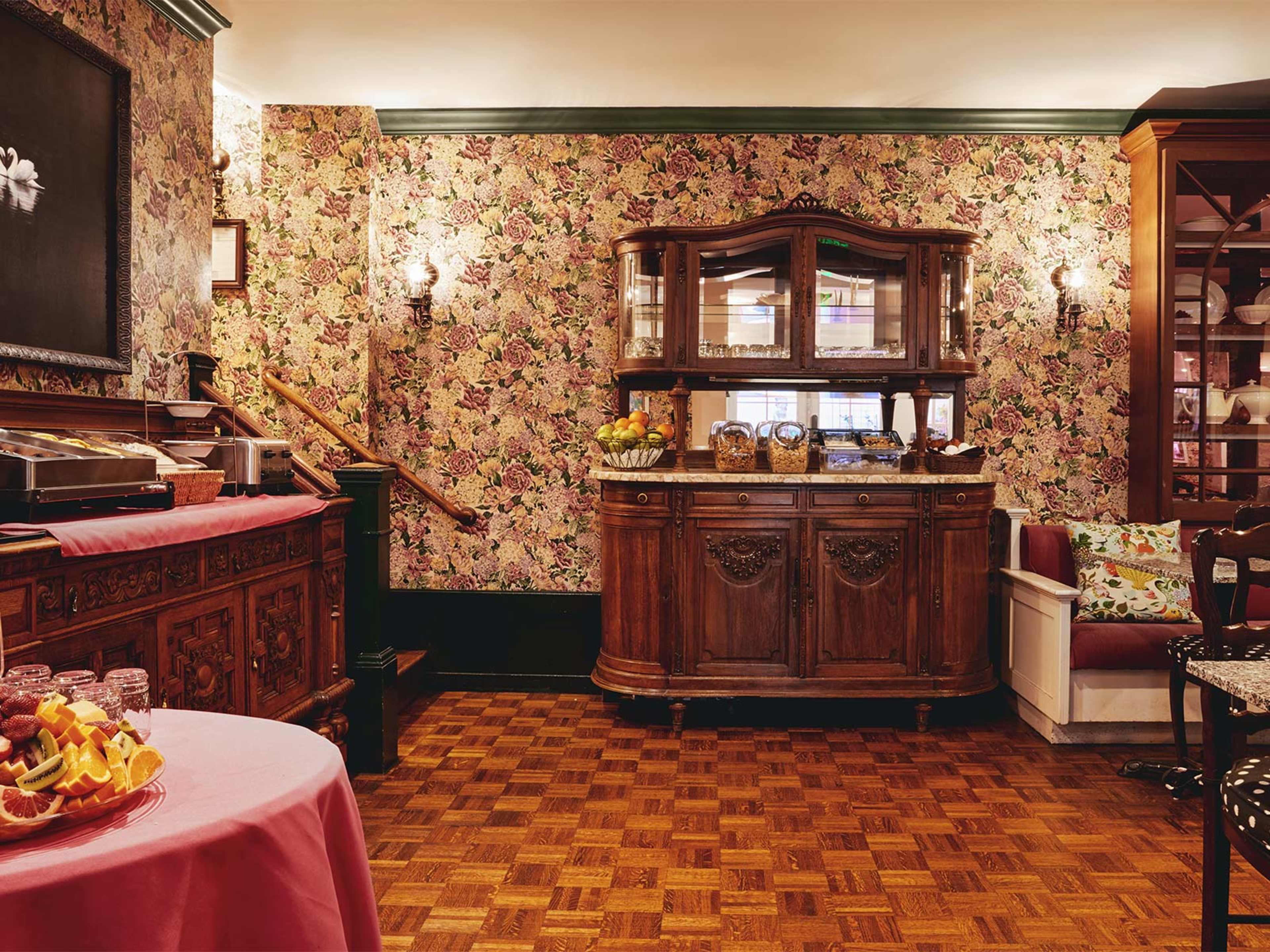 A vintage-style dining area with floral wallpaper, a wooden sideboard, and a small table set with fruit.