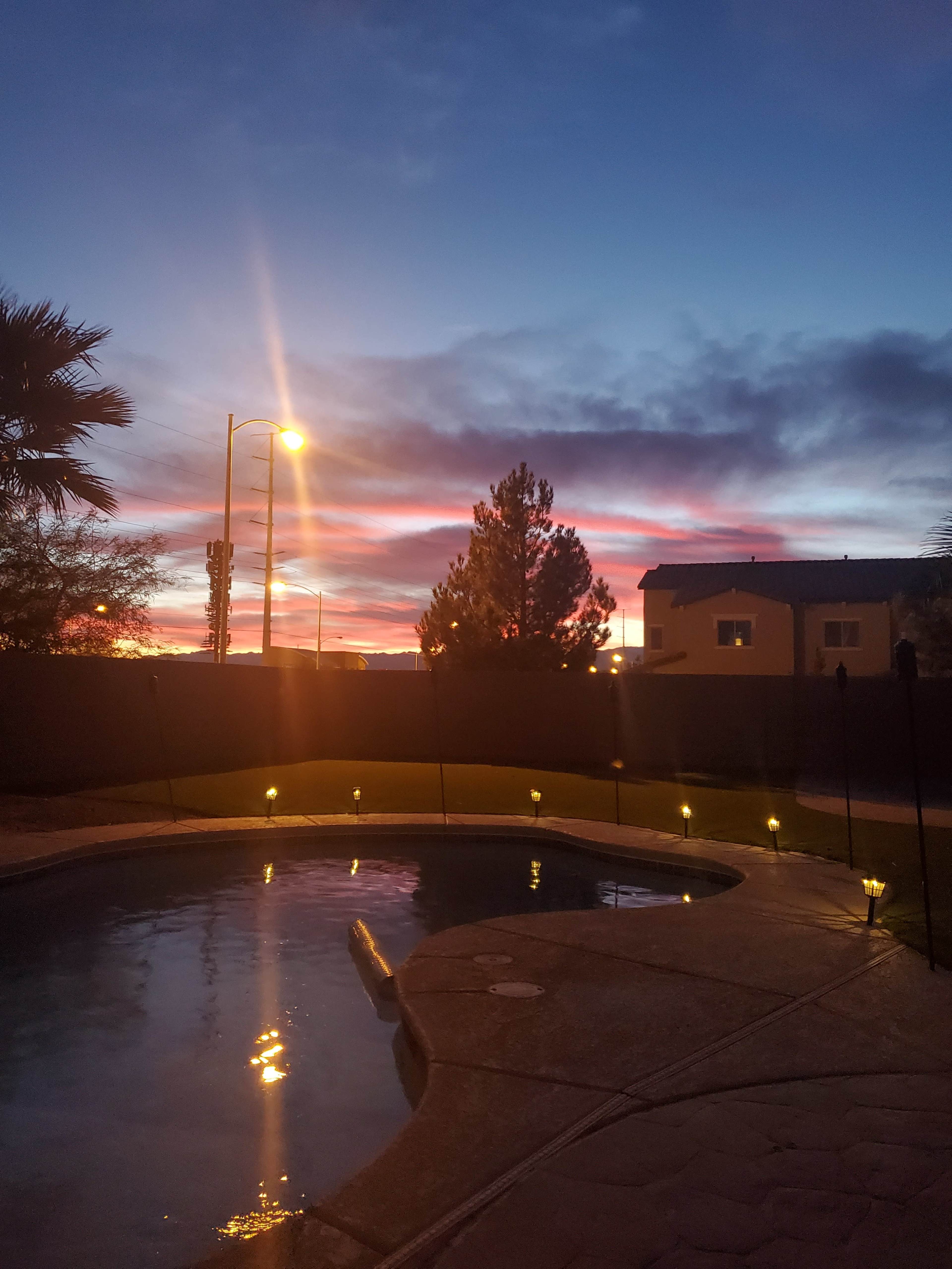 A swimming pool reflects the colorful sunset sky, with a few lights illuminating the surrounding area.