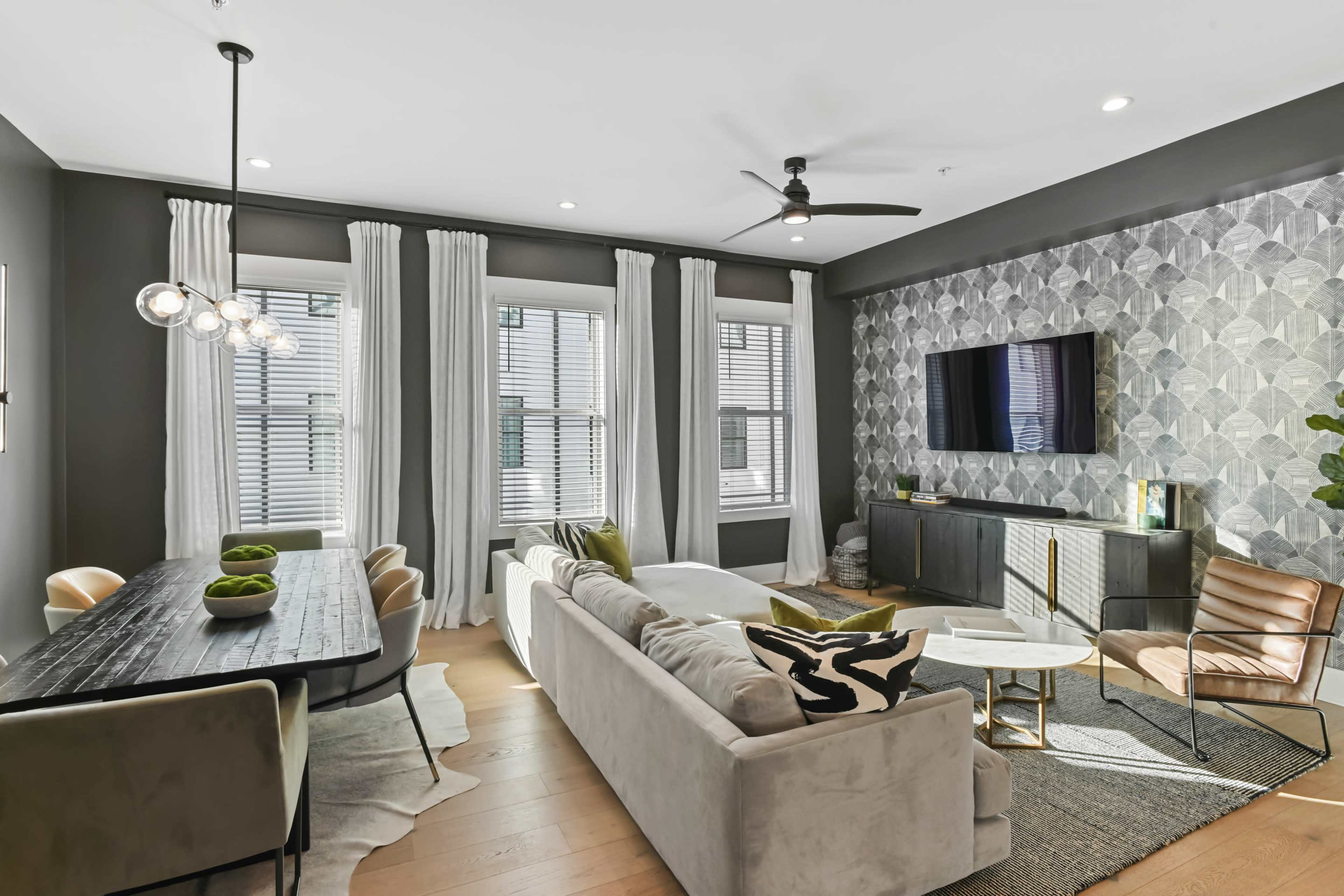 The image shows a modern living room and dining area with a gray color scheme, featuring a sectional sofa, a dining table, large windows with curtains, and a patterned accent wall.
