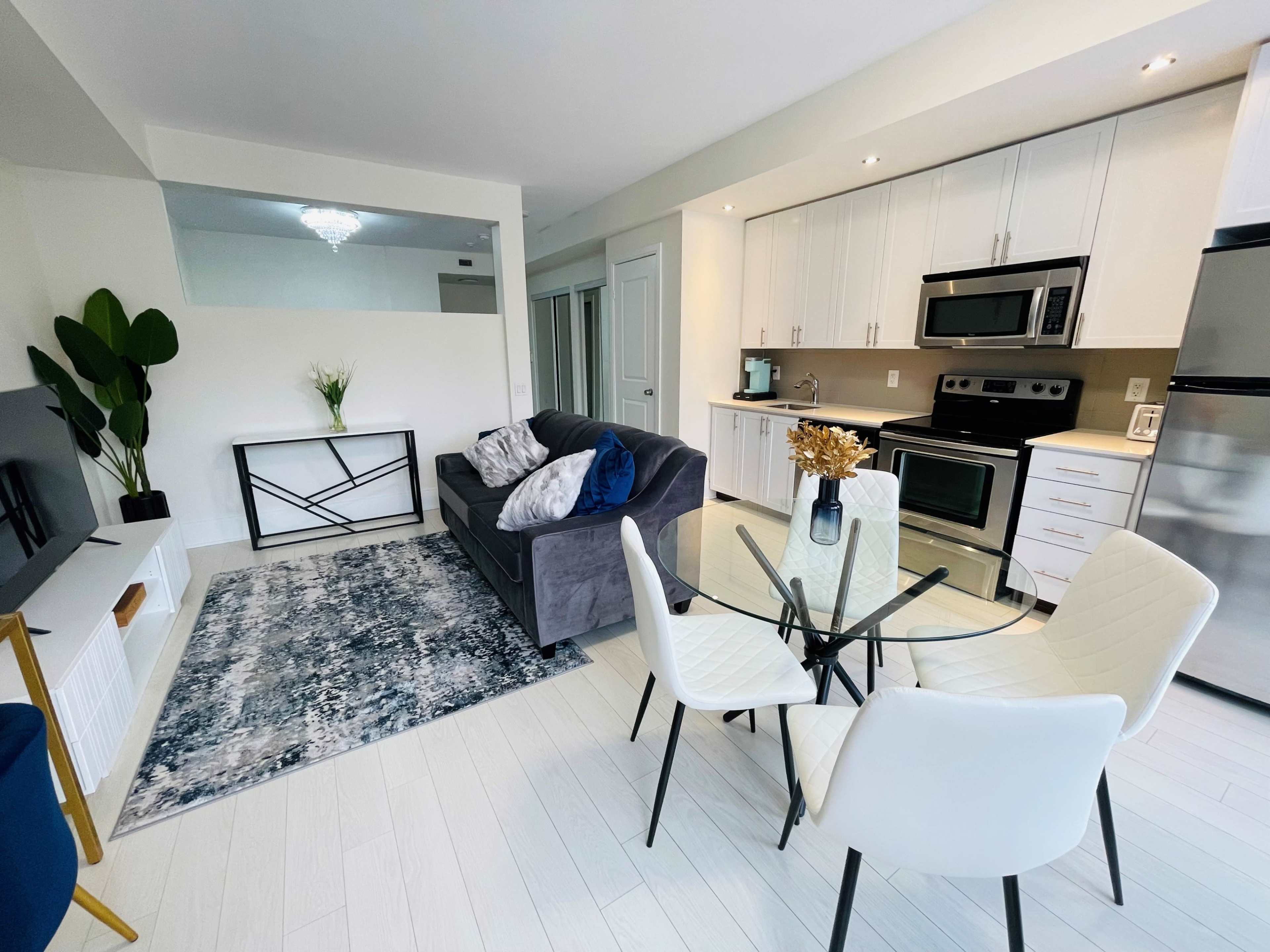 A modern living area featuring a gray sofa, a glass dining table with four chairs, and kitchen appliances in a bright space.