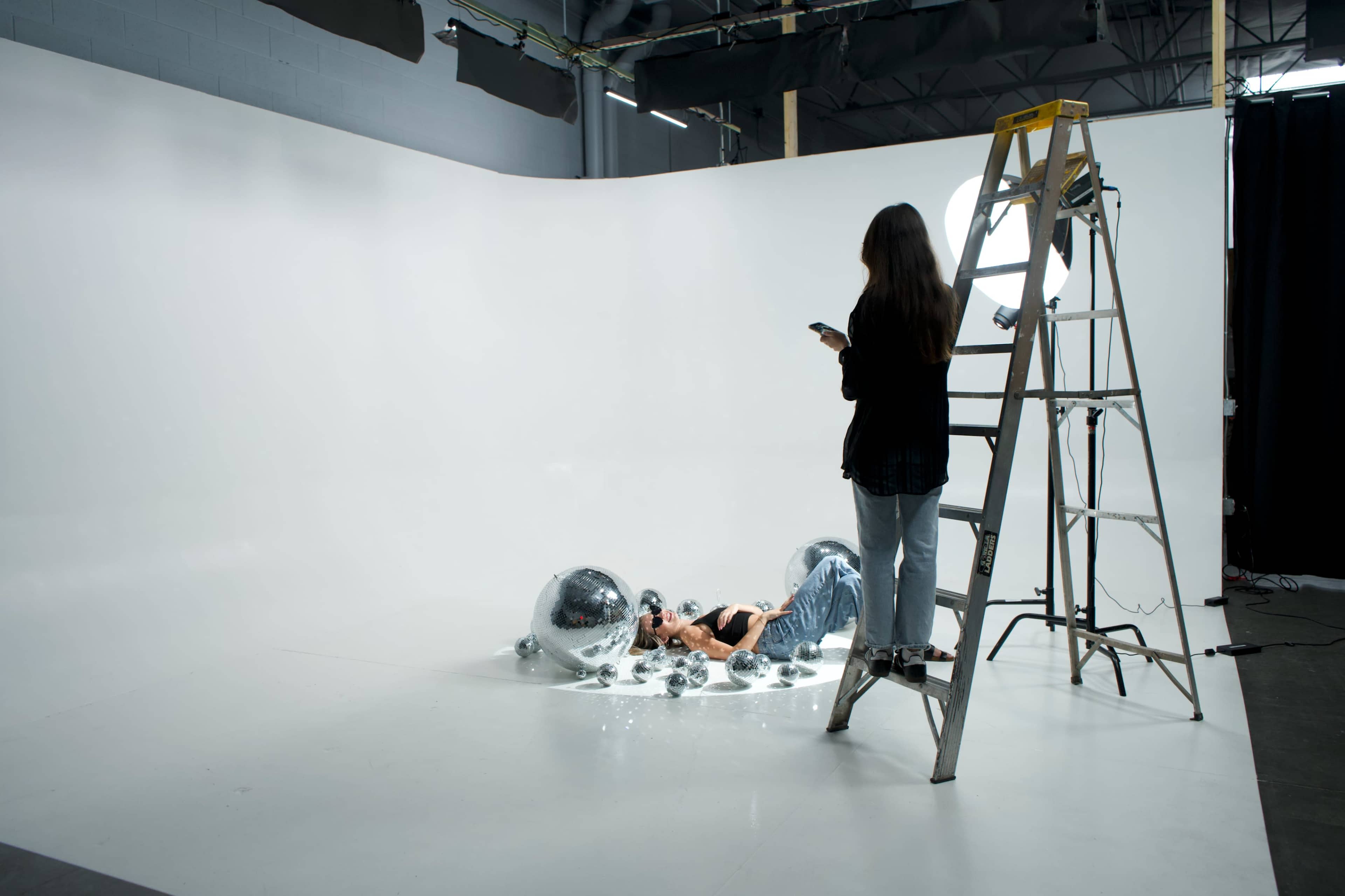 A person lies on the floor surrounded by large reflective spheres, while another person stands on a ladder holding a phone in a studio with a plain white backdrop.