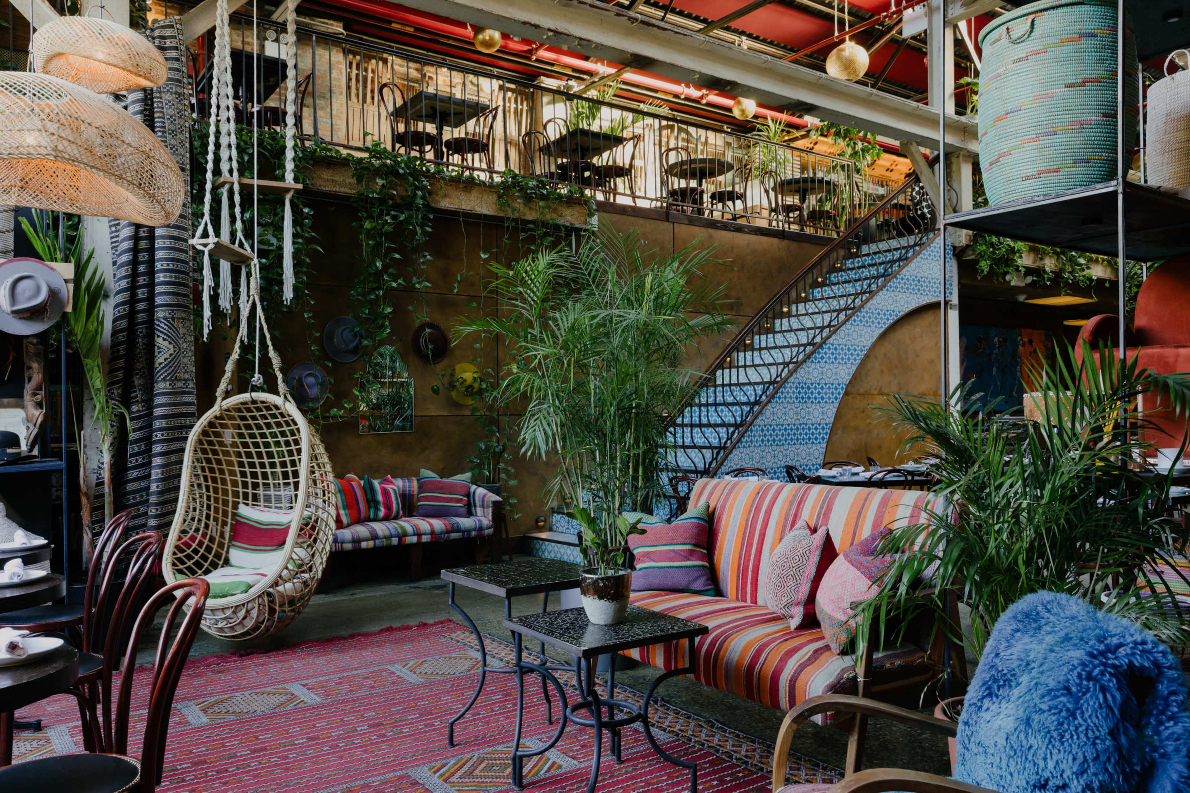 A vibrant indoor café featuring colorful striped seating, hanging plants, a decorative staircase, and a cozy atmosphere with a mix of plants and furnishings.