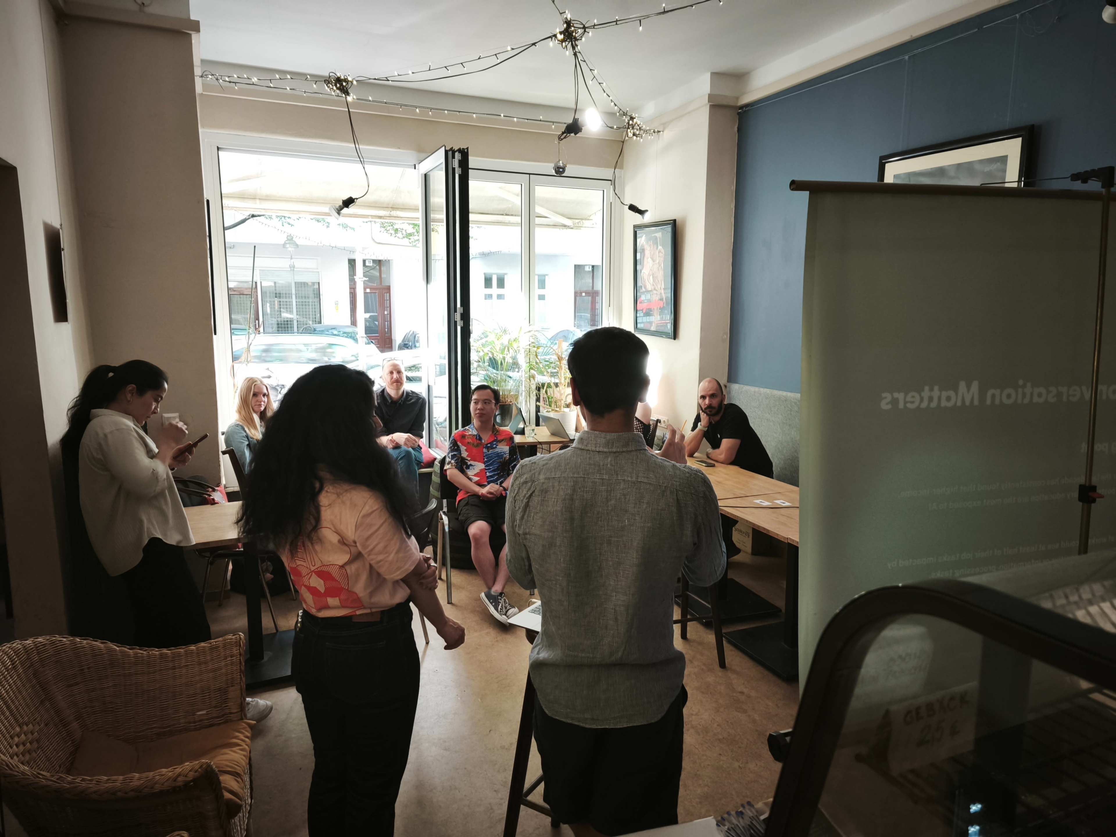 A person stands at the front of a café while a small group of people seated at tables engages attentively.