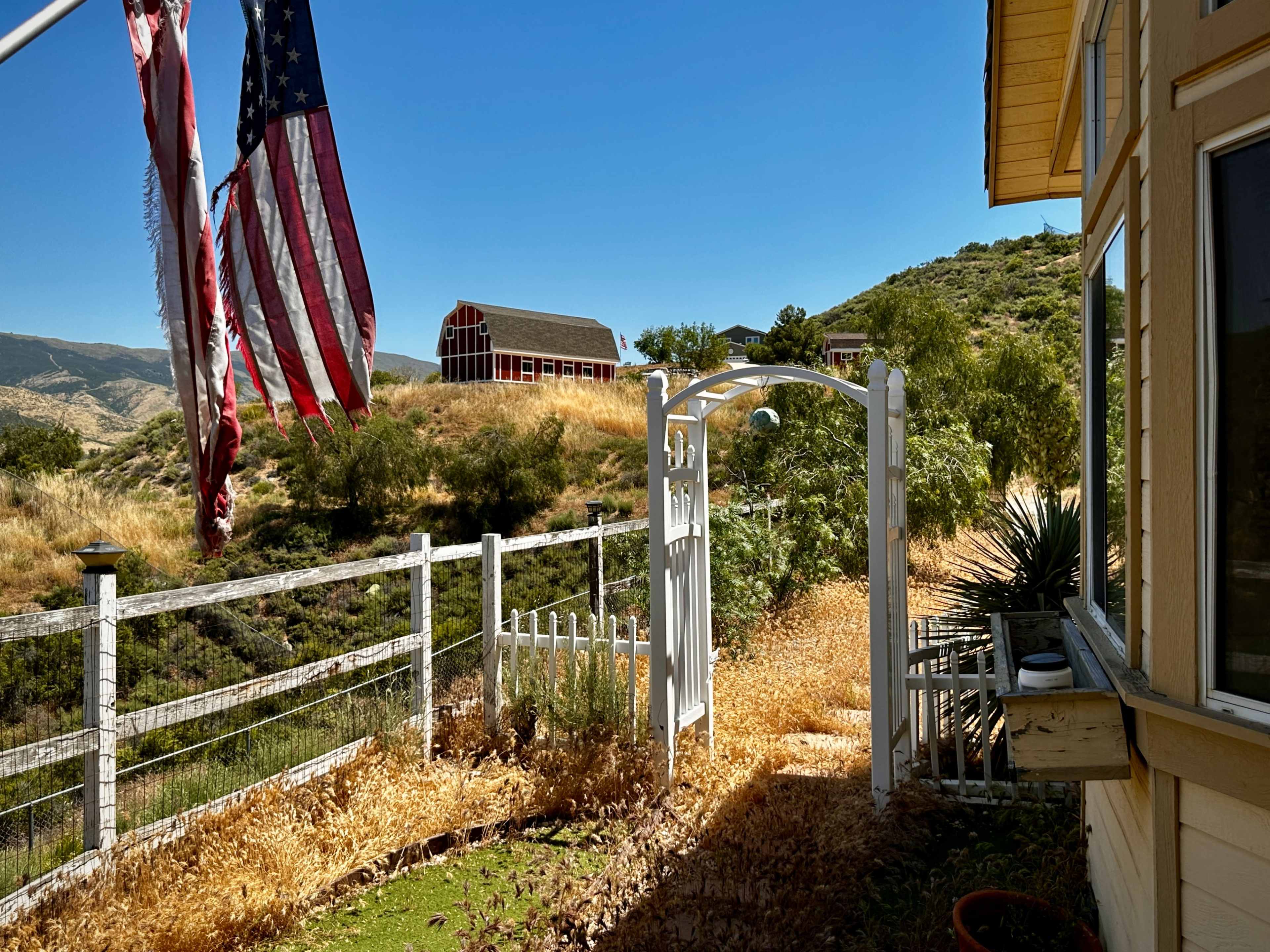 A white gate leading to a pathway bordered by a fence, with American flags flying on the left and a red barn on a hillside in the background.