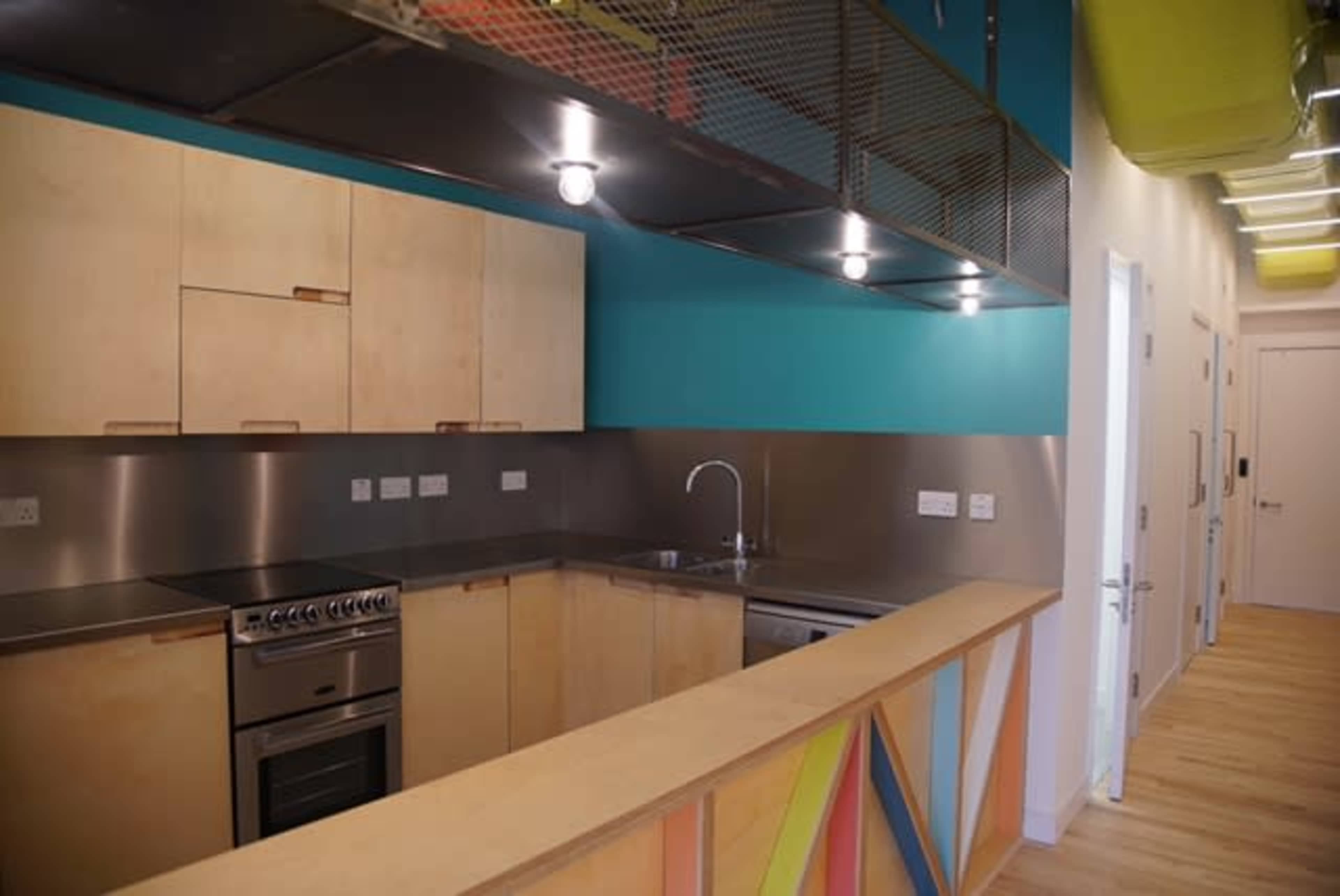 A modern kitchen with wooden cabinetry, stainless steel appliances, and a bright blue wall, with a view of a hallway beyond.
