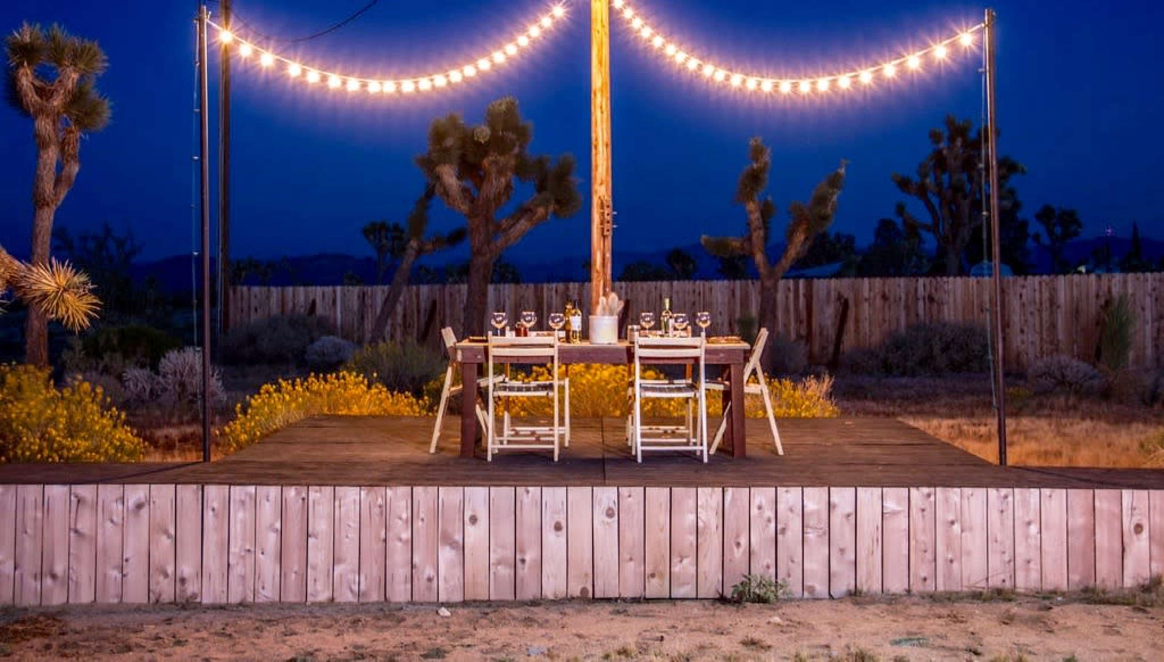 A dining table with chairs is set up on a wooden platform under string lights in a desert landscape at night.