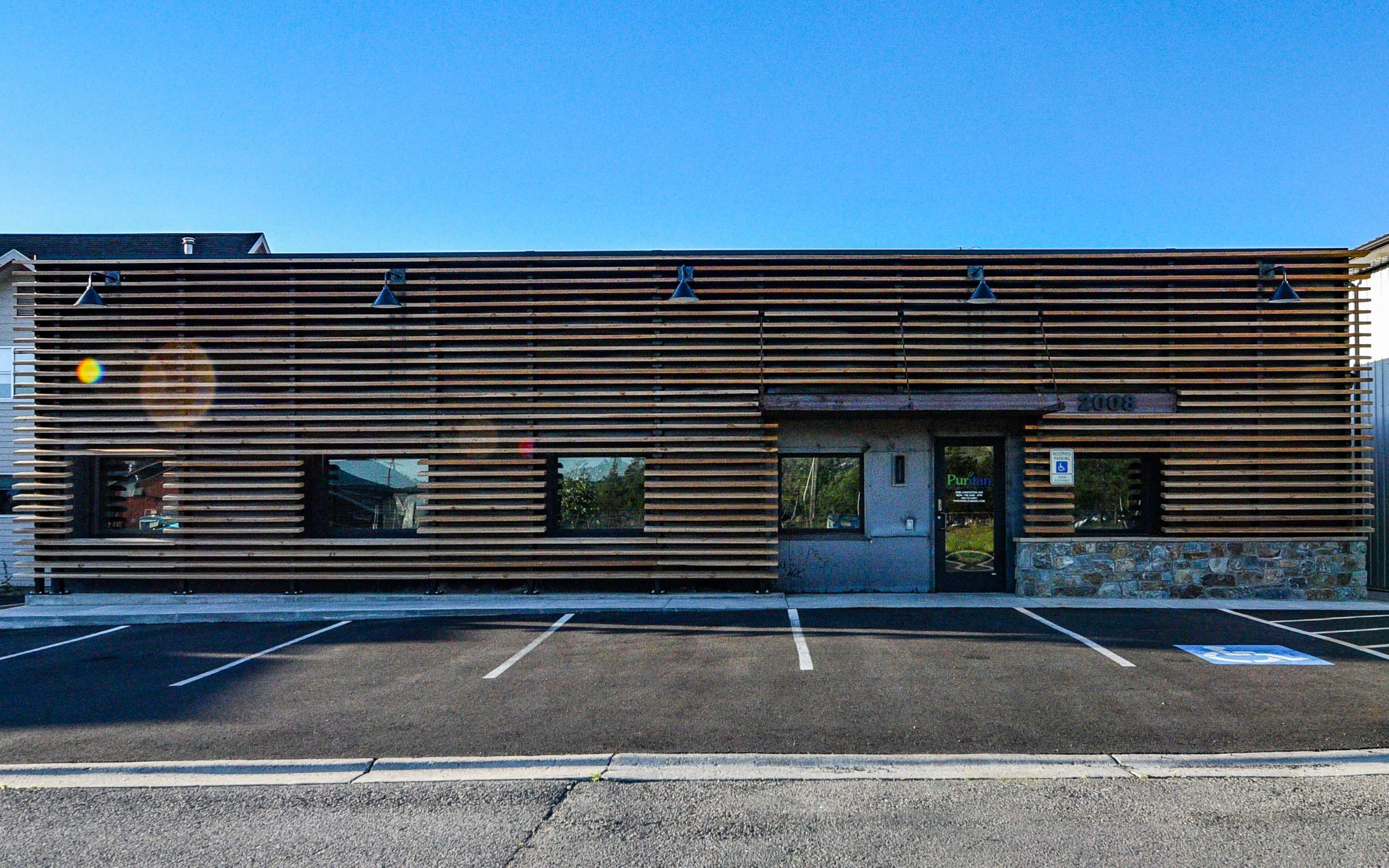 A building with horizontal wooden slats and a stone base, featuring windows and a single entrance, situated in a paved parking lot.