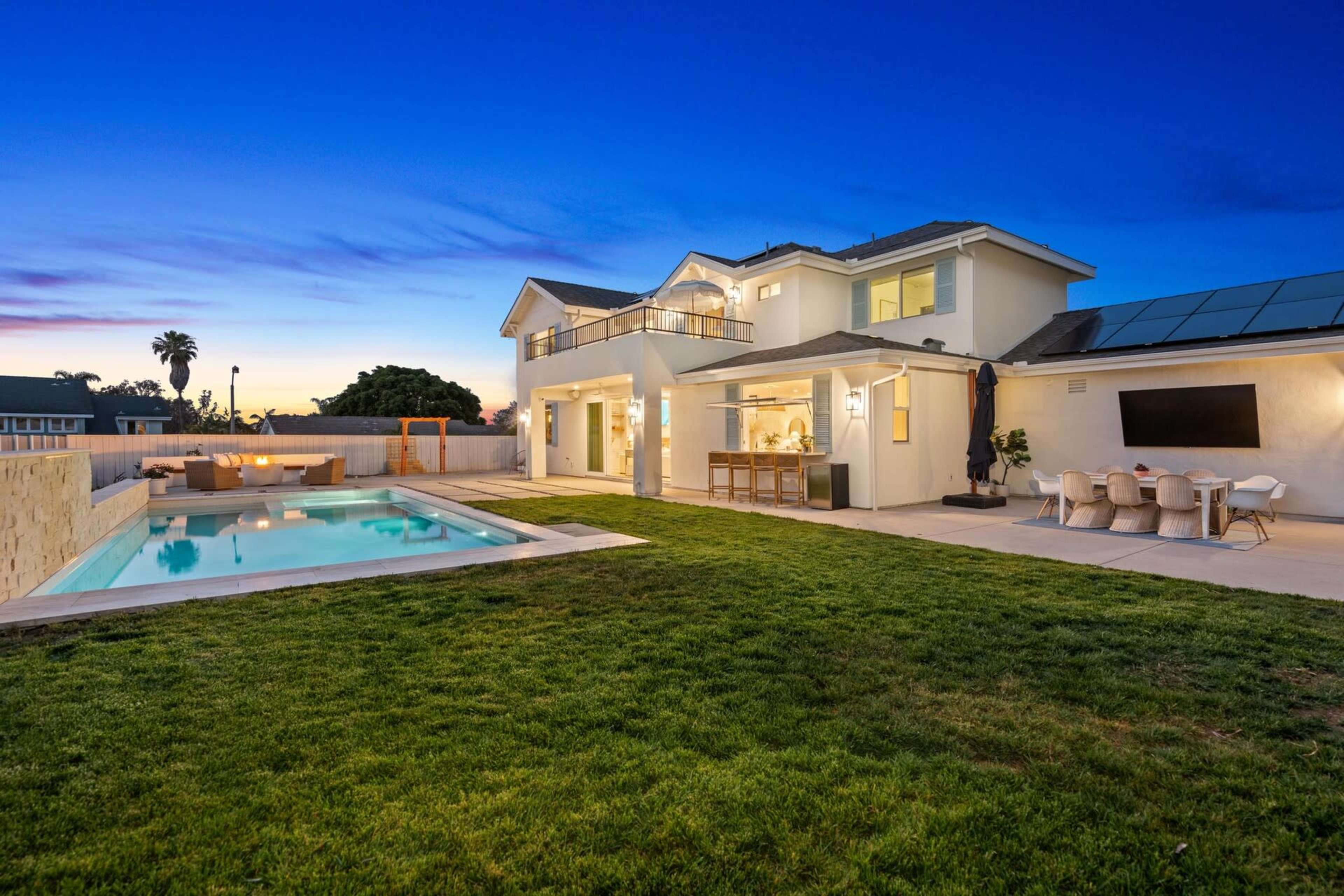 A modern two-story house features an outdoor pool and a spacious patio area with seating, set against a twilight sky.