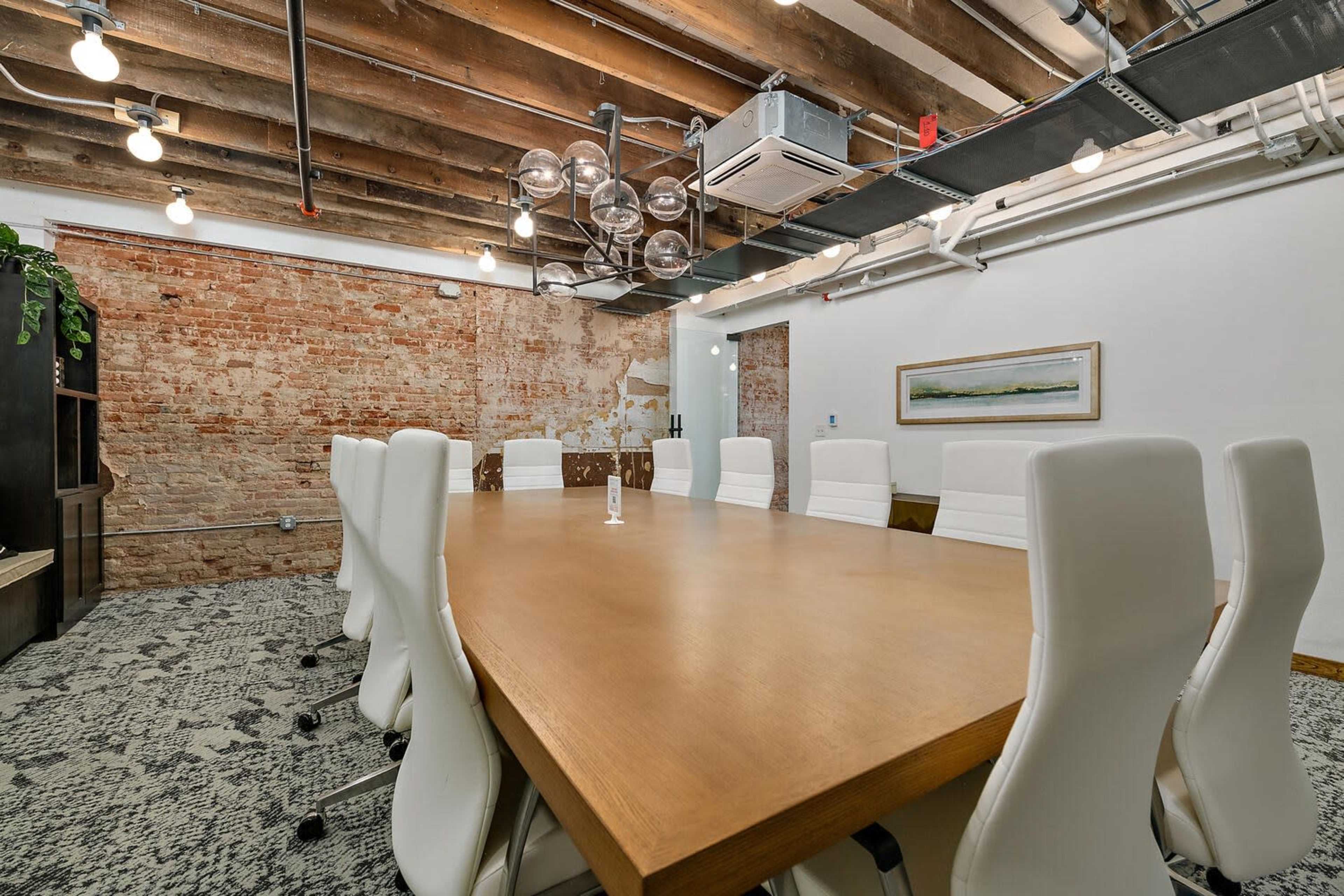 A spacious conference room features a large wooden table surrounded by white chairs, with exposed brick walls and modern lighting fixtures.