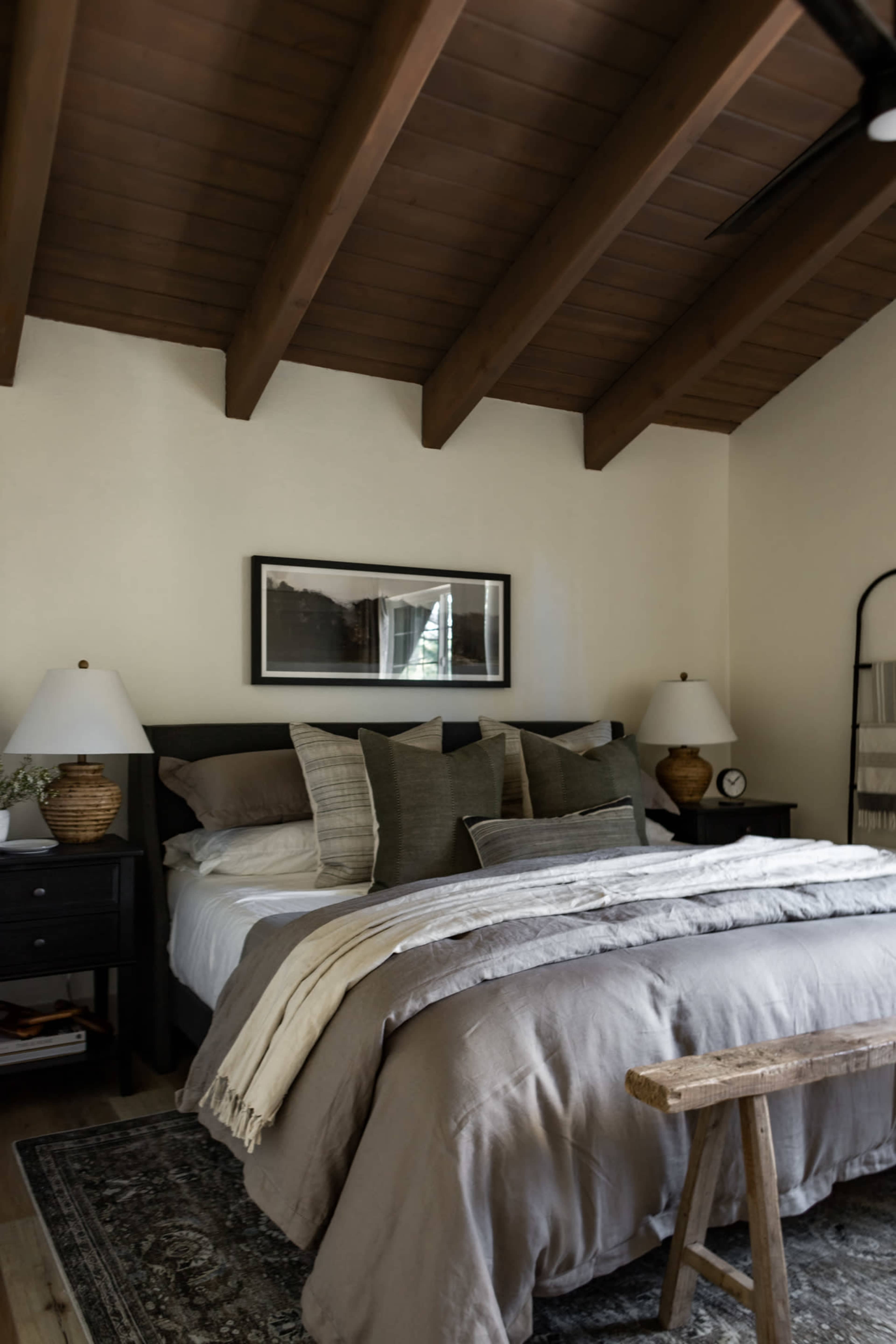 The image shows a cozy bedroom featuring a king-sized bed with neutral bedding, wooden beams on the ceiling, and two bedside lamps on matching nightstands.