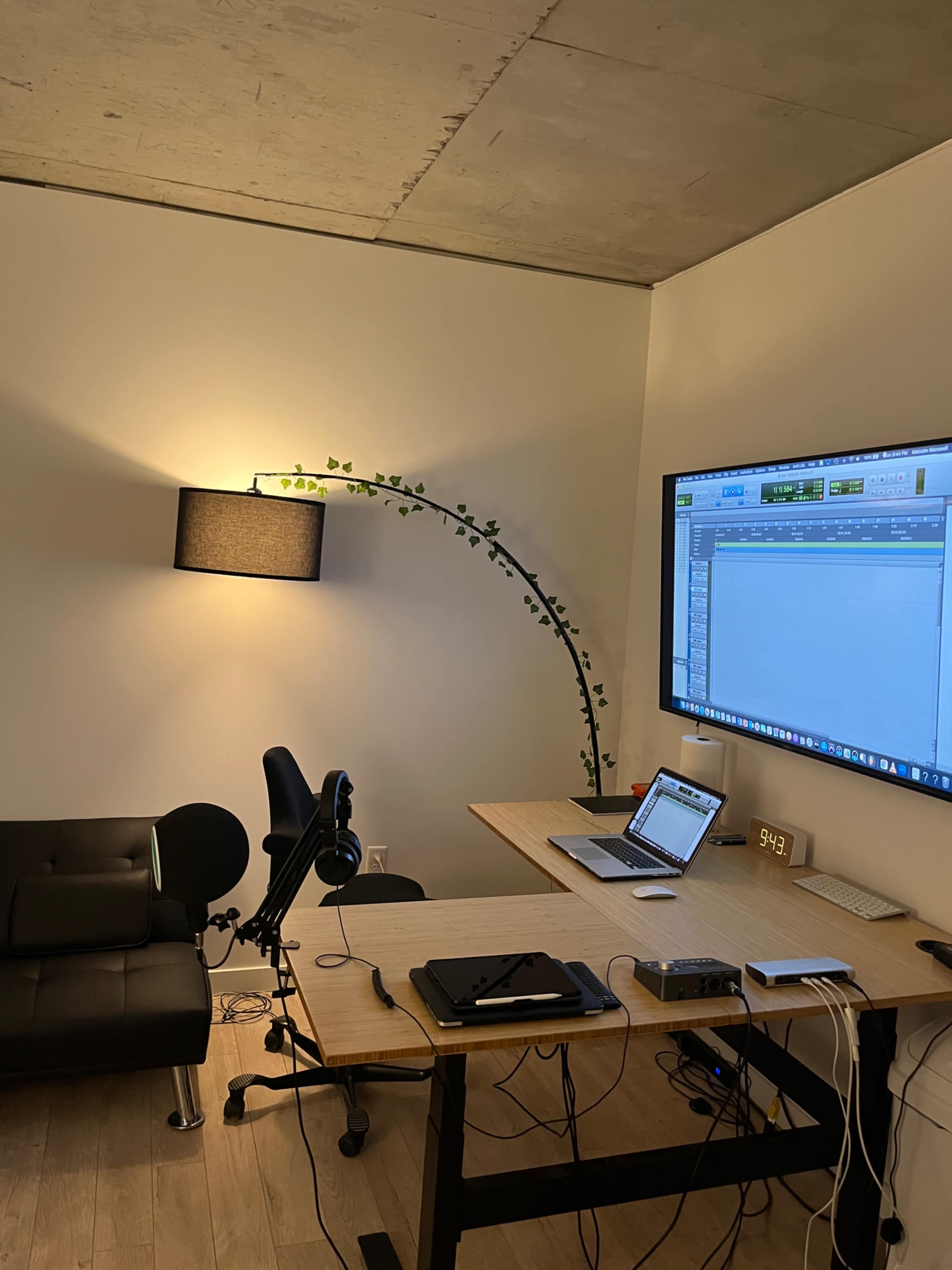 A modern workspace features a large monitor displaying software, a laptop, a microphone, and a comfortable couch under an arching lamp with greenery.