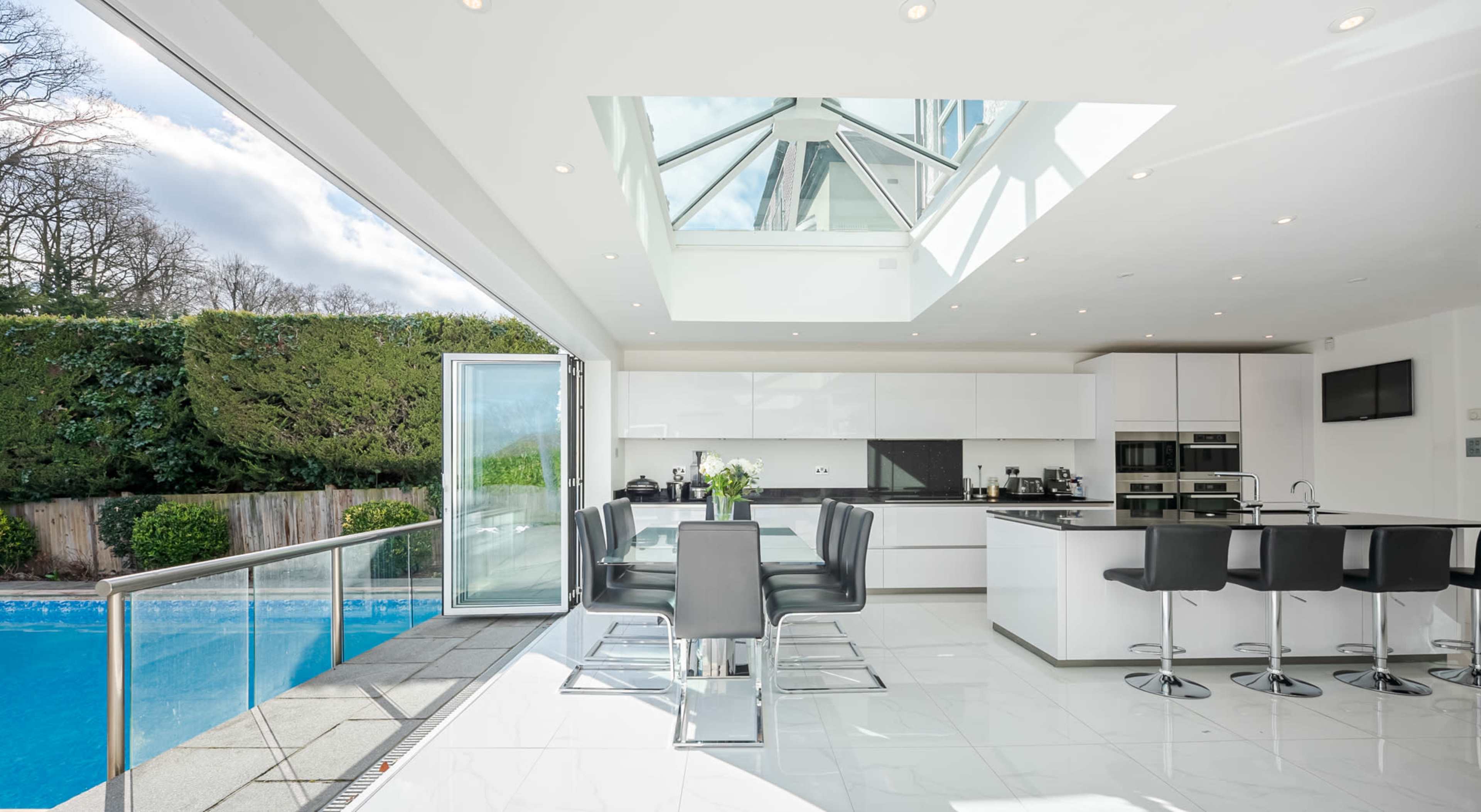 A modern kitchen with a glass ceiling, featuring sleek white cabinets, a dining table set for six, and a view of a patio with a swimming pool.