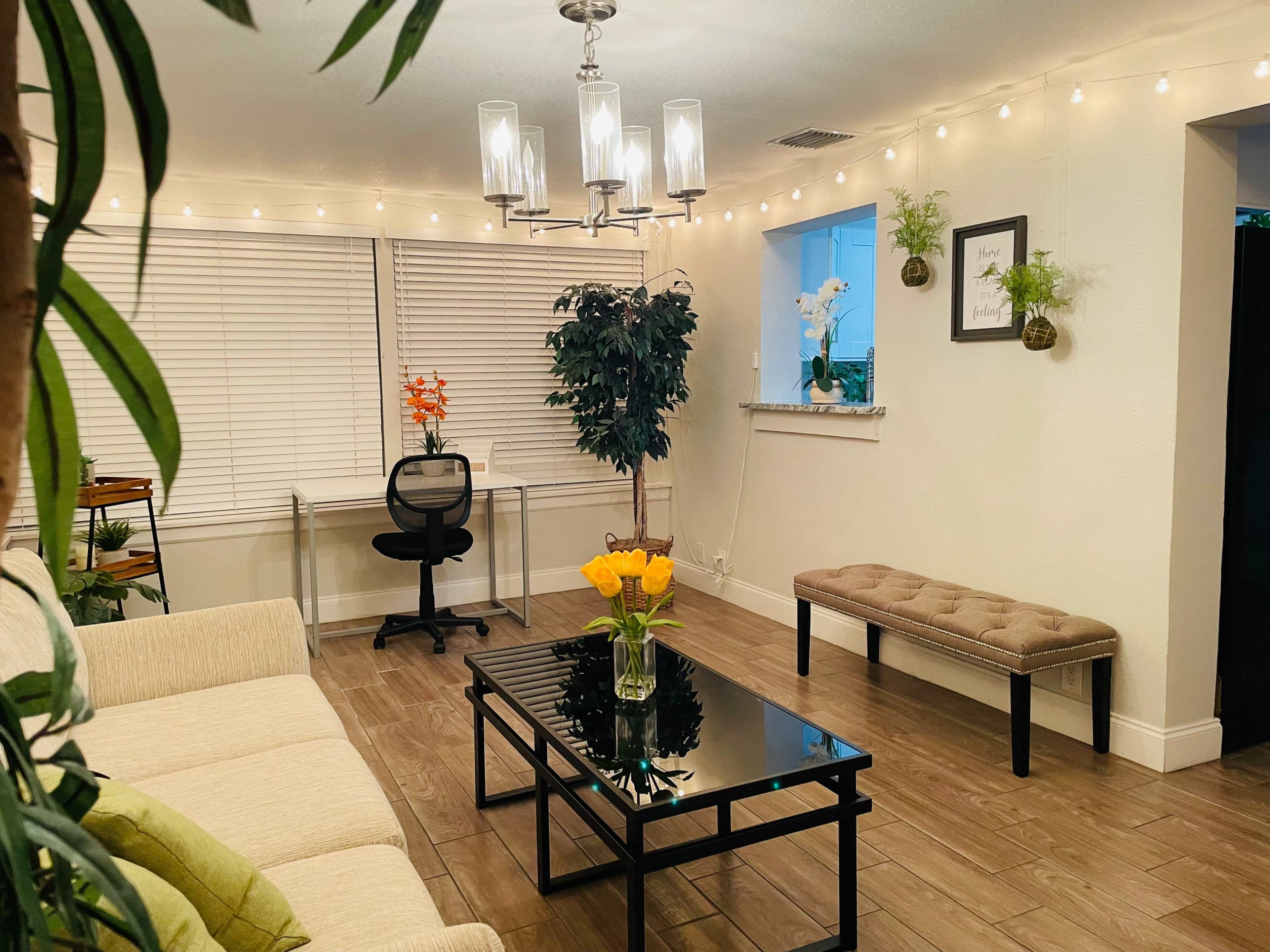 A well-lit living room features a light-colored sofa, a glass coffee table with a vase of orange flowers, and a decorative plant, with a desk and chair in the background.