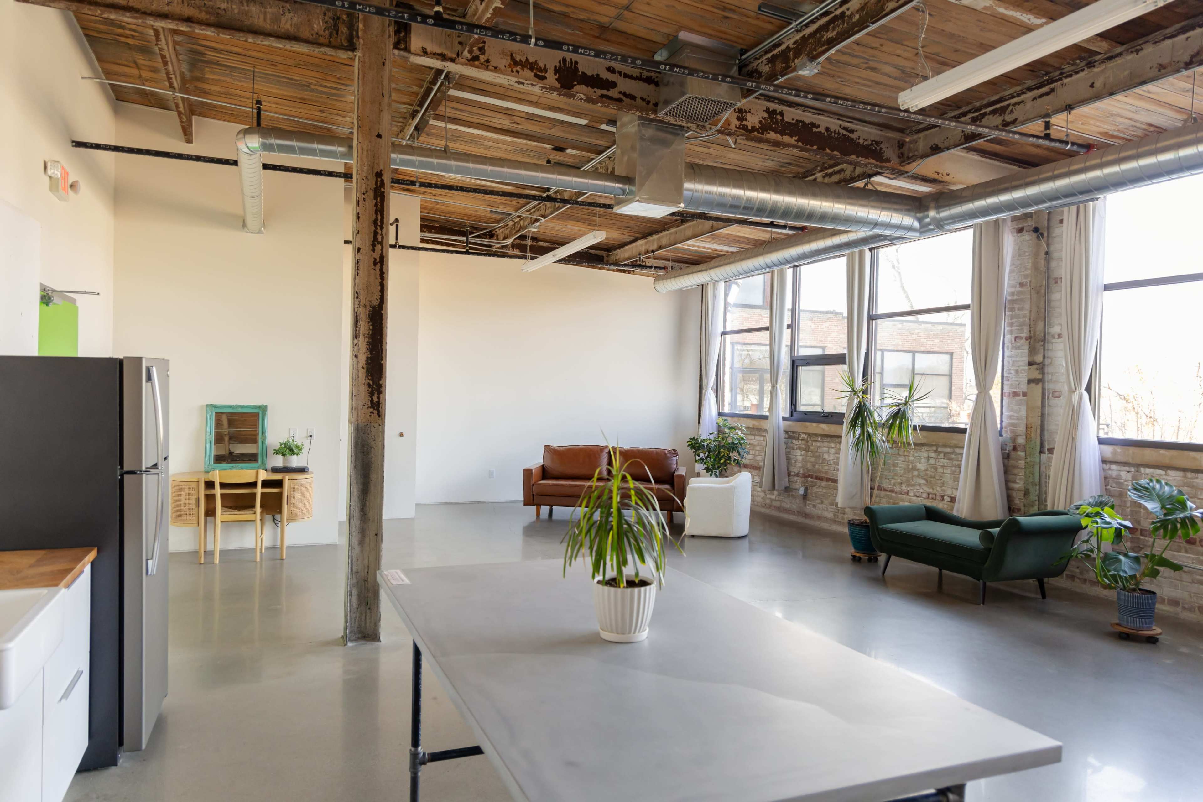 A spacious, industrial-style room with large windows, concrete flooring, wooden beams, and a mix of furniture including a sofa and chairs.