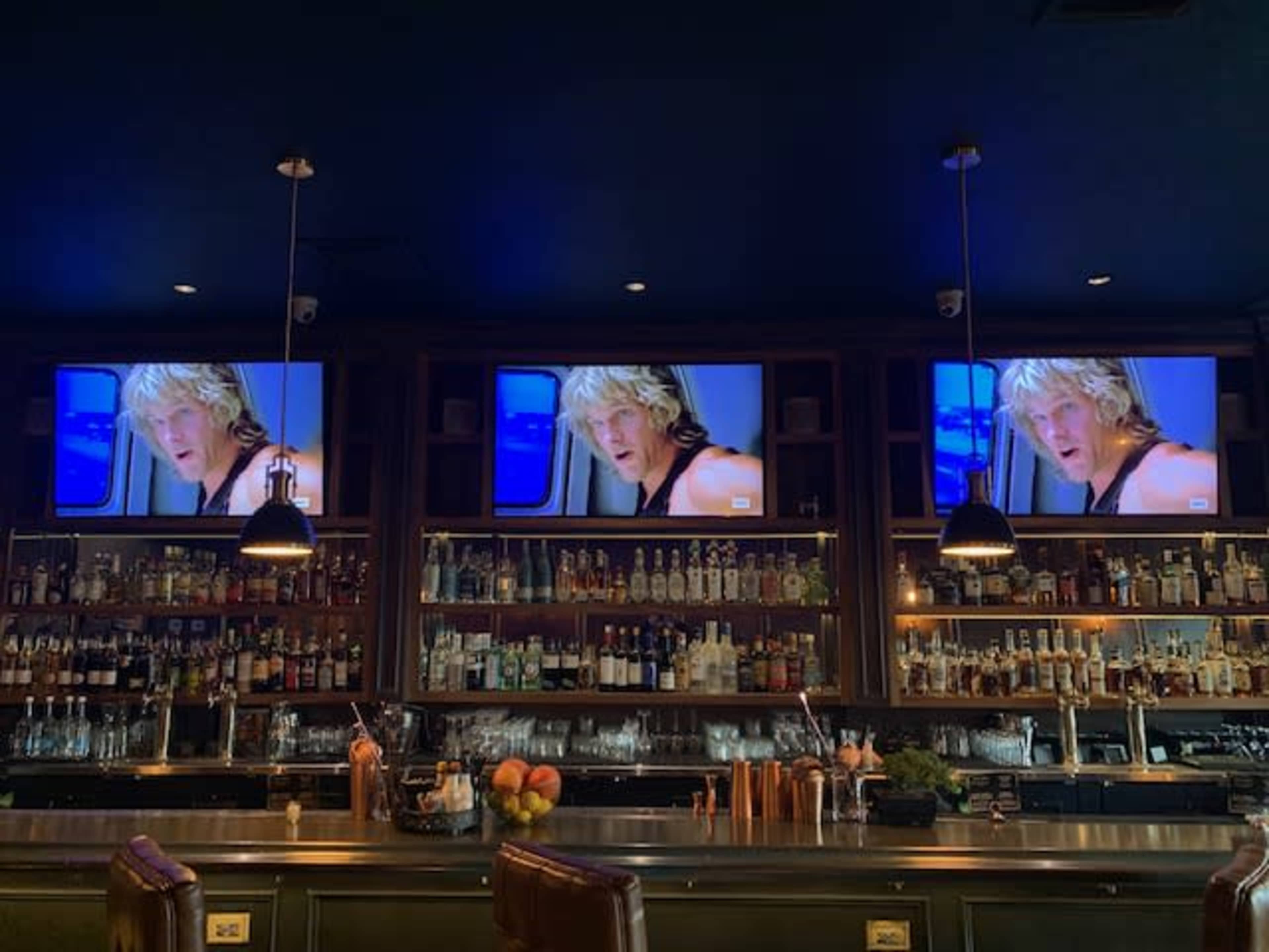 The image shows a bar with three televisions mounted above a well-stocked shelf of liquor bottles, displaying a scene from a movie featuring a man with blonde hair.